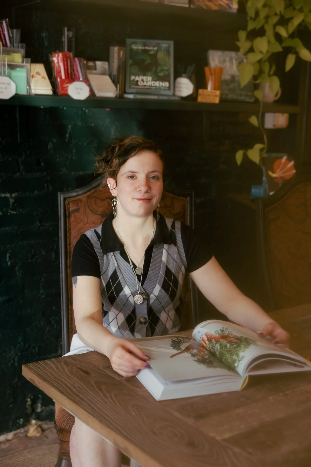 A young woman with short, curly brown hair and earrings is sitting at a wooden table, looking at an open magazine with images of nature, including trees. She is wearing a black, gray, and white argyle sweater vest over a black shirt and has a slight 