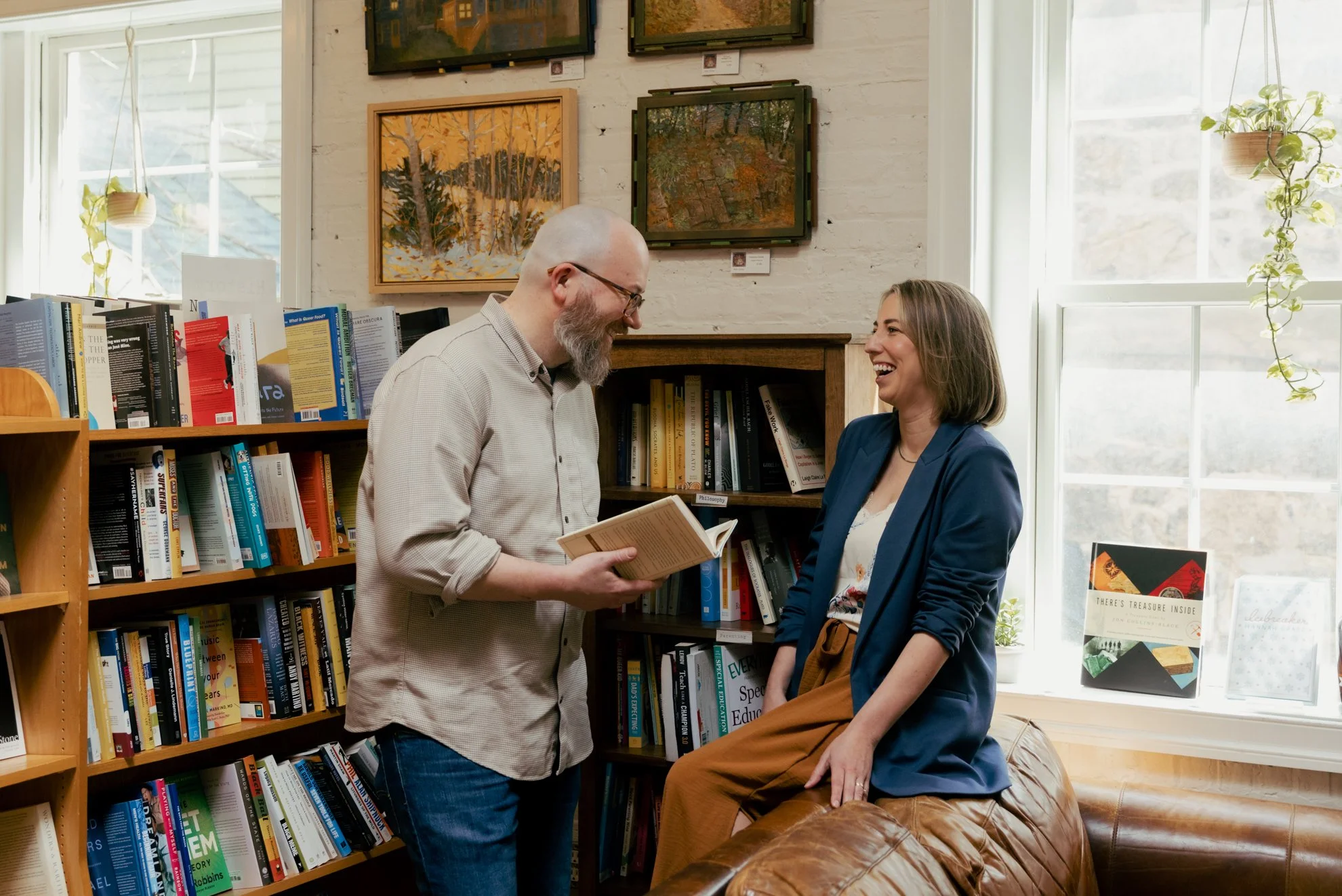 A man with a beard and glasses stands holding a book, smiling at a woman who is sitting on a brown leather couch. The woman has shoulder-length hair and is wearing a blue blazer. They are in a cozy bookstore or library, with bookshelves filled with b