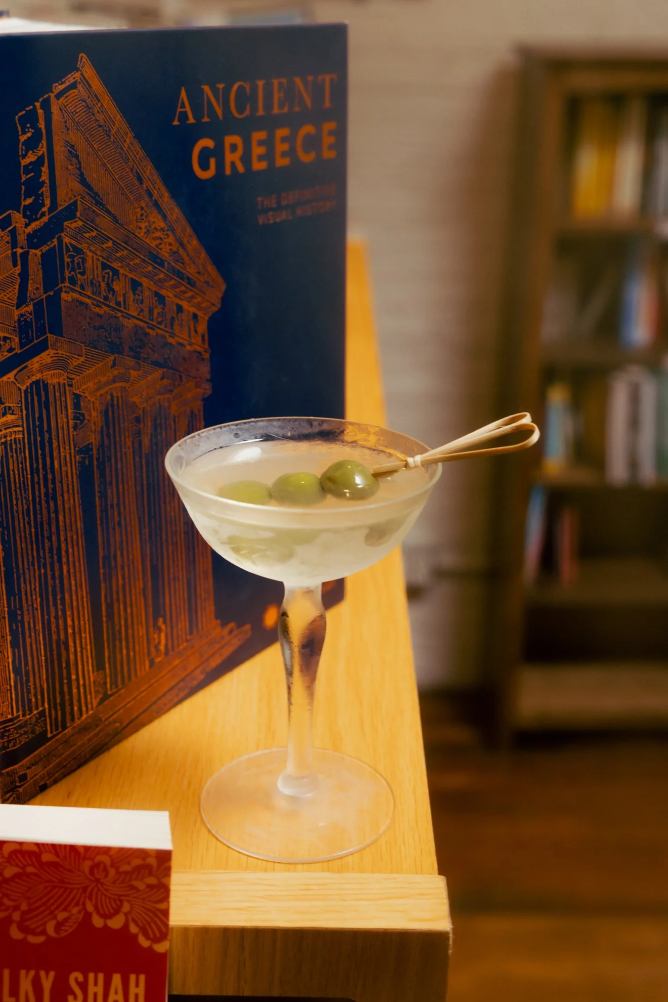 A cocktail glass with green olives, placed on a wooden surface, with a blue book titled 'Ancient Greece' in the background.