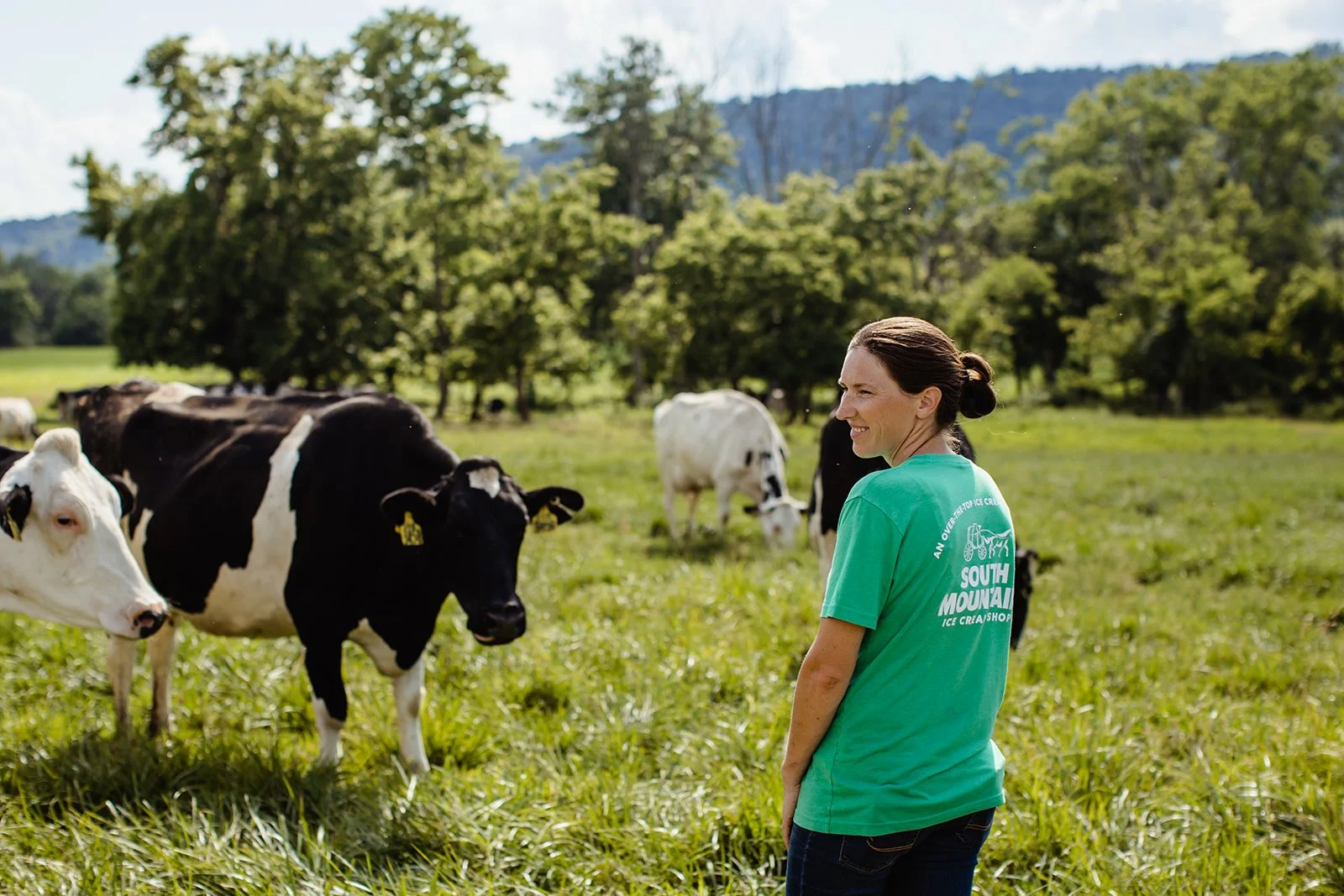 A woman standing in a grassy field with black and white cows, trees, and mountains in the background on a sunny day.