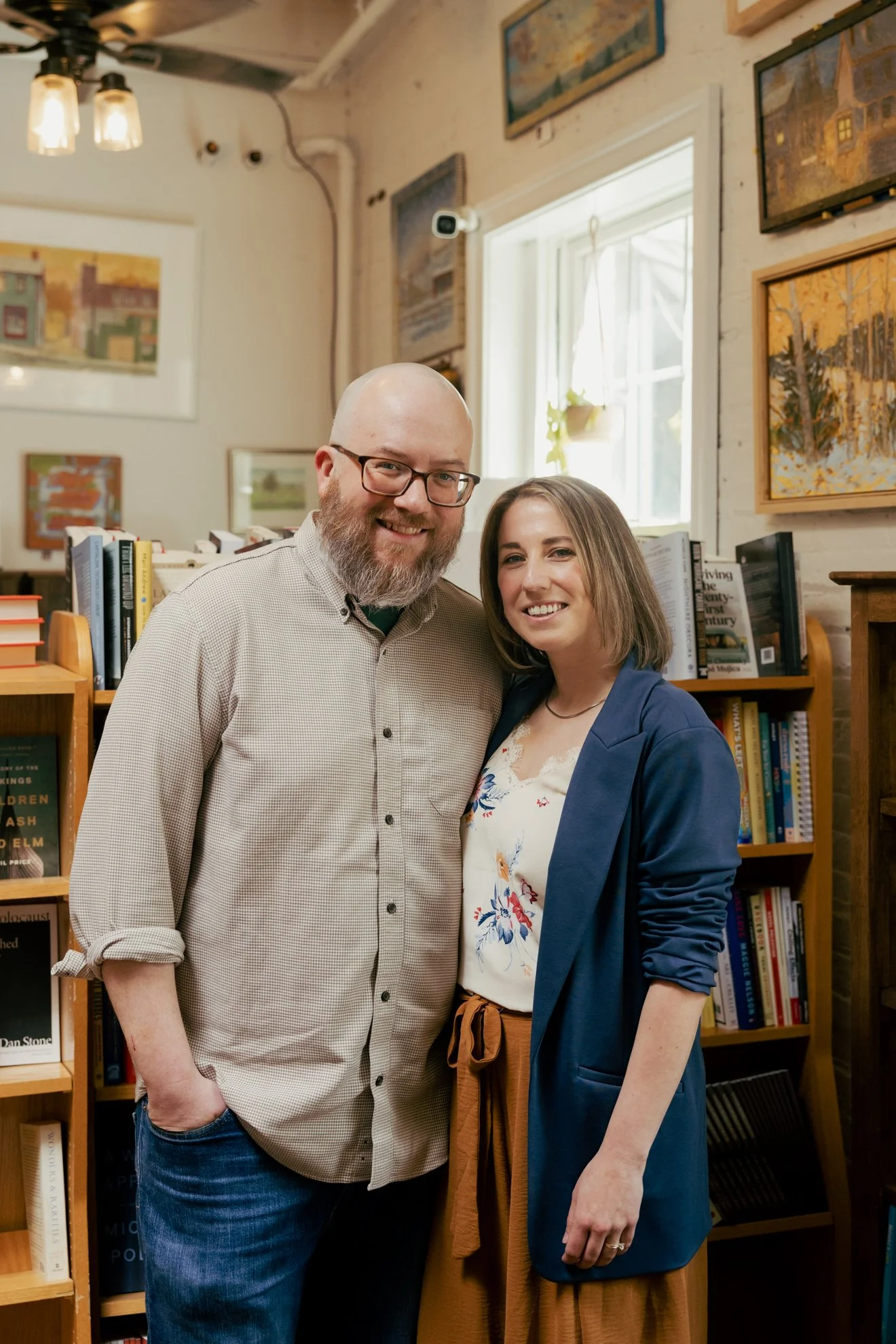 A man with glasses, a beard, and a checked shirt standing next to a woman with shoulder-length hair, wearing a blue blazer and a floral blouse, inside a room with bookshelves and framed art on the walls.