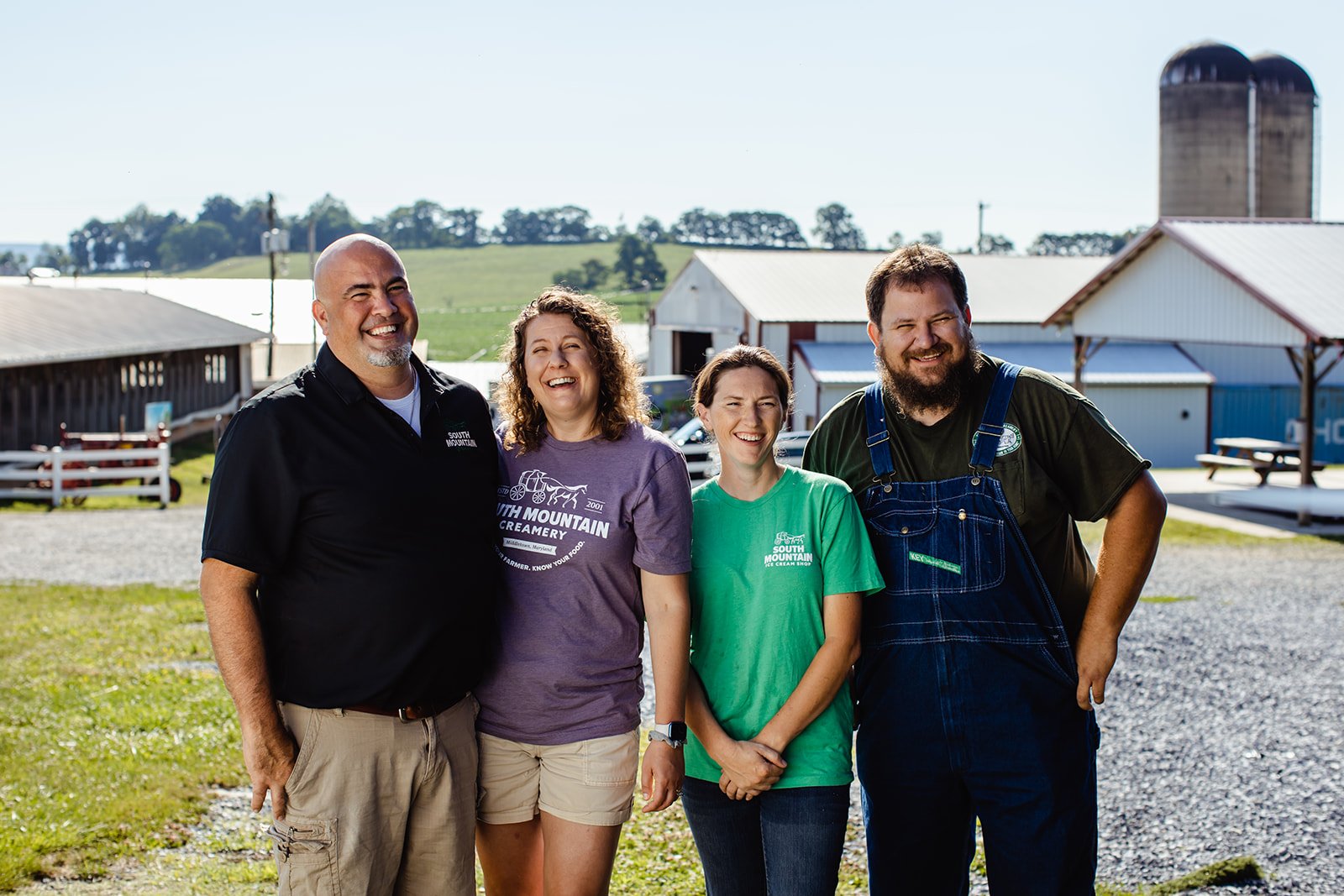Four smiling people standing outdoors in front of farm buildings, dressed casually with some wearing shirts with farm branding, enjoying a sunny day.