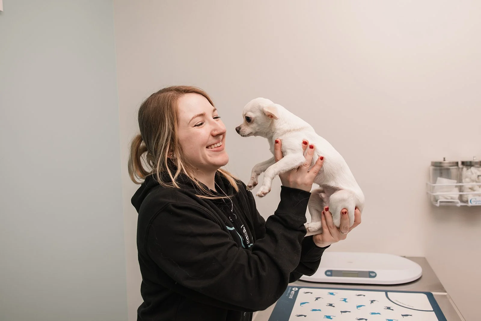 A woman with reddish hair smiling and holding a small white puppy in her hands in a veterinary office or clinic.