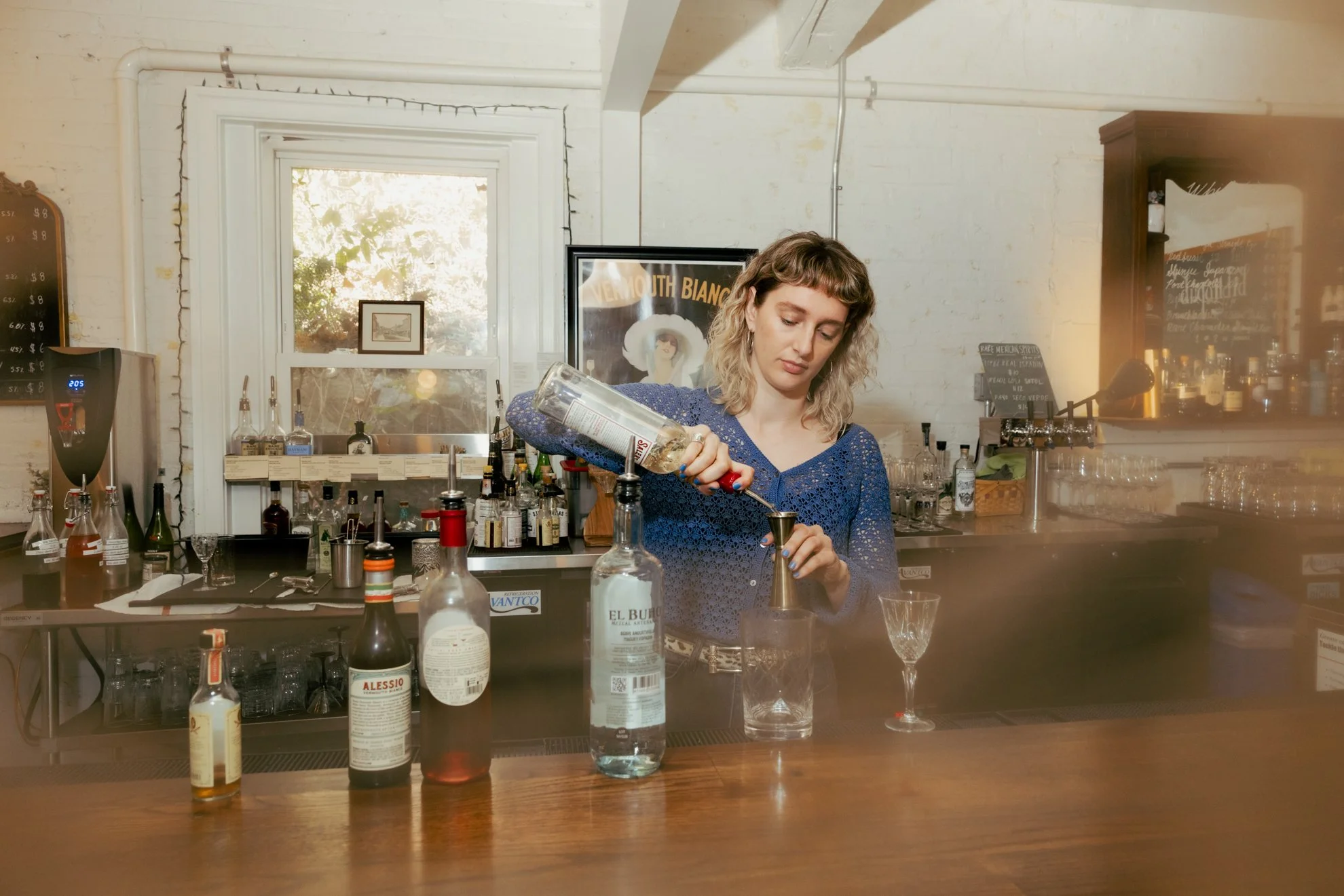 A woman in a blue dress is preparing a cocktail at a bar counter. The bar area has various bottles of alcohol and bar tools, with a window behind her letting in natural light. The setting appears cozy and vintage, with artwork and a chalkboard menu o