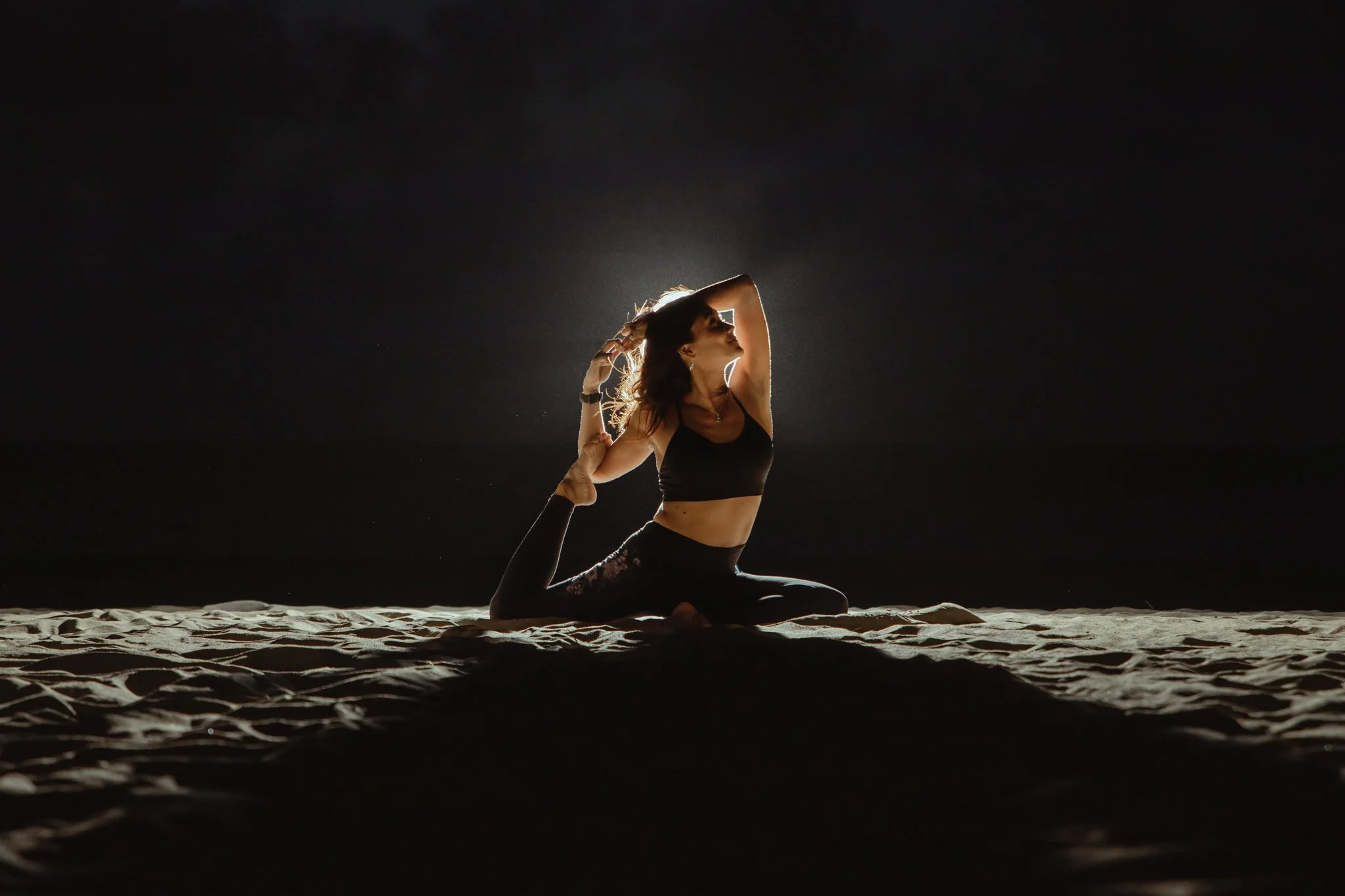 A woman practicing yoga on the sand at night, performing a backbend pose with her arm over her head, illuminated from behind creating a silhouette against a dark sky.