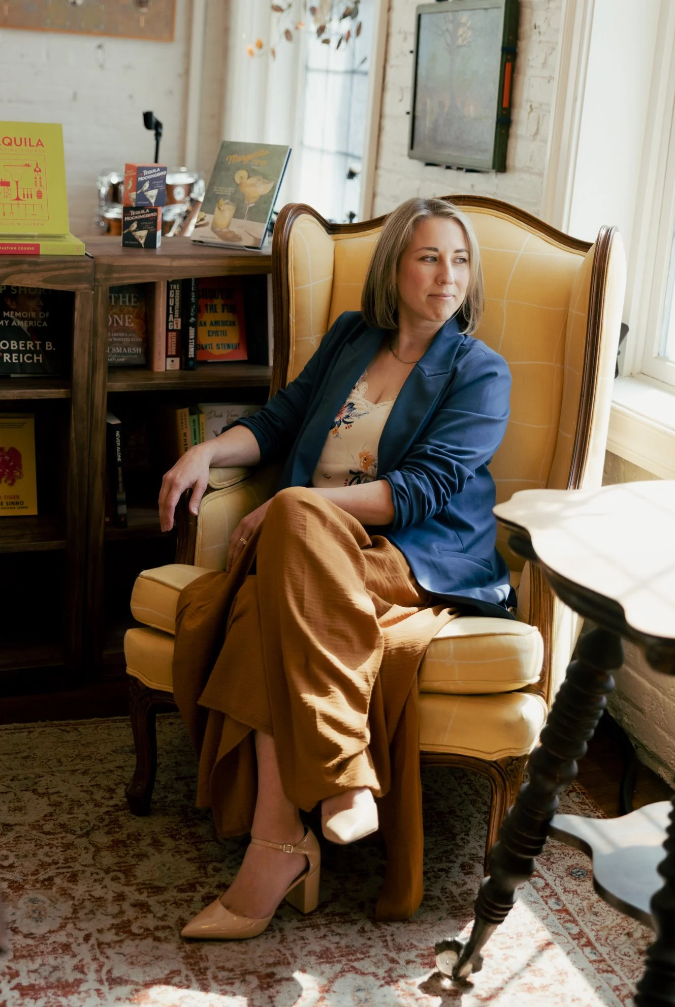 Woman sitting in a vintage yellow armchair near a window, looking to her right with a contemplative expression. She has shoulder-length light brown hair and is dressed in a blue blazer over a floral top, tan pants, and beige high heels. Behind her is
