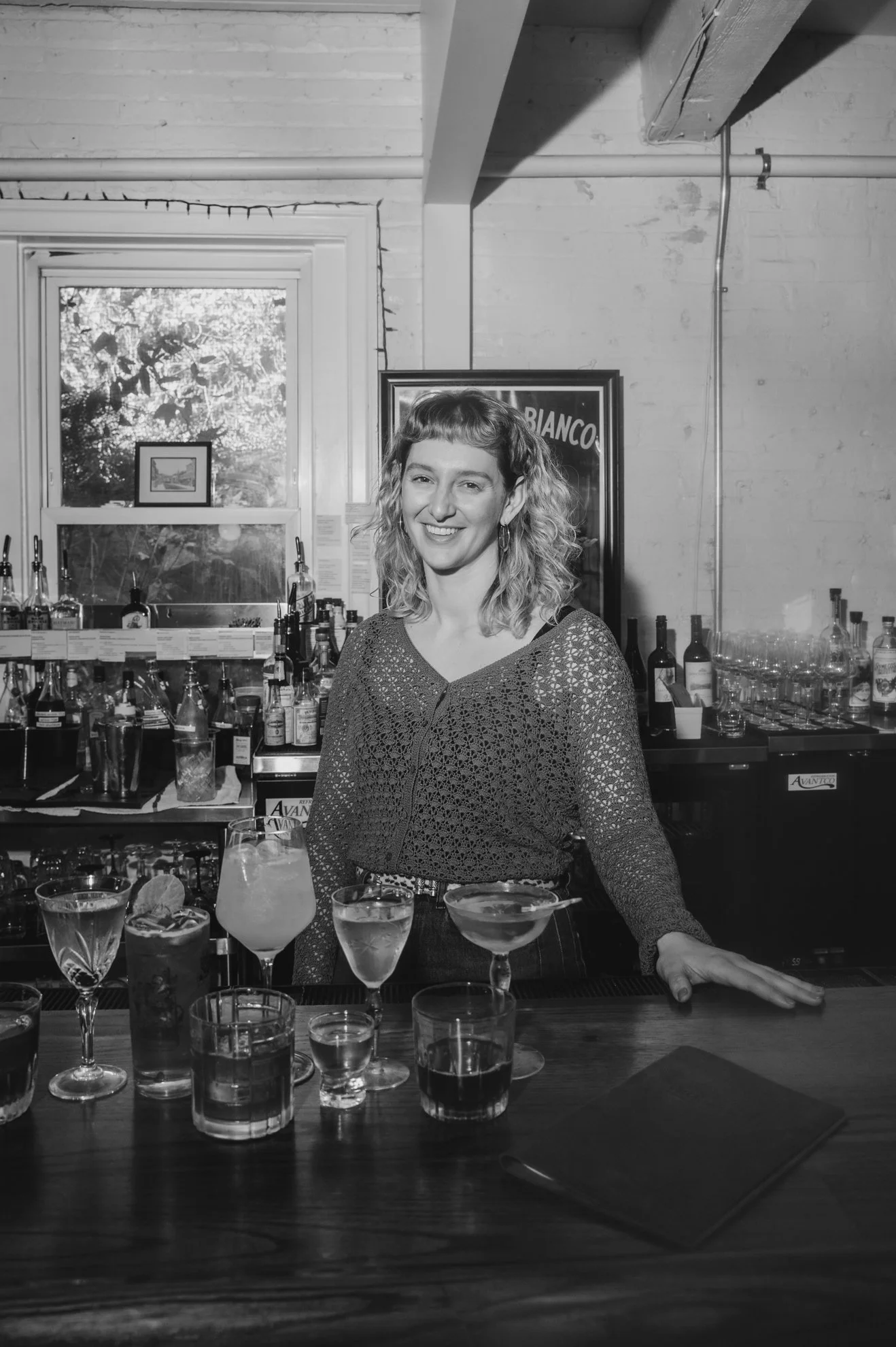 A woman behind a bar with a selection of cocktails on a wooden counter in a well-lit room with a window. The woman is smiling and has curly hair, wearing a patterned blouse.