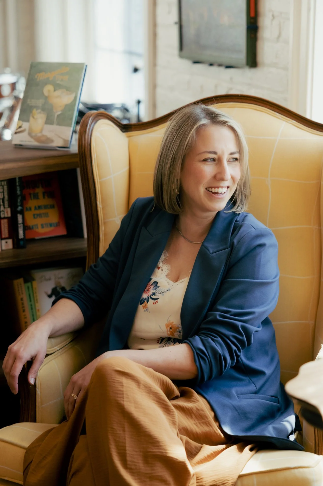 A woman with shoulder-length blond hair, smiling and sitting on a yellow sofa in a cozy, well-lit room with books and a television in the background.