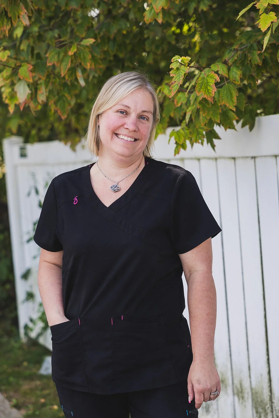A smiling woman with short blonde hair wearing black scrubs, standing outdoors in front of a white fence and green foliage.