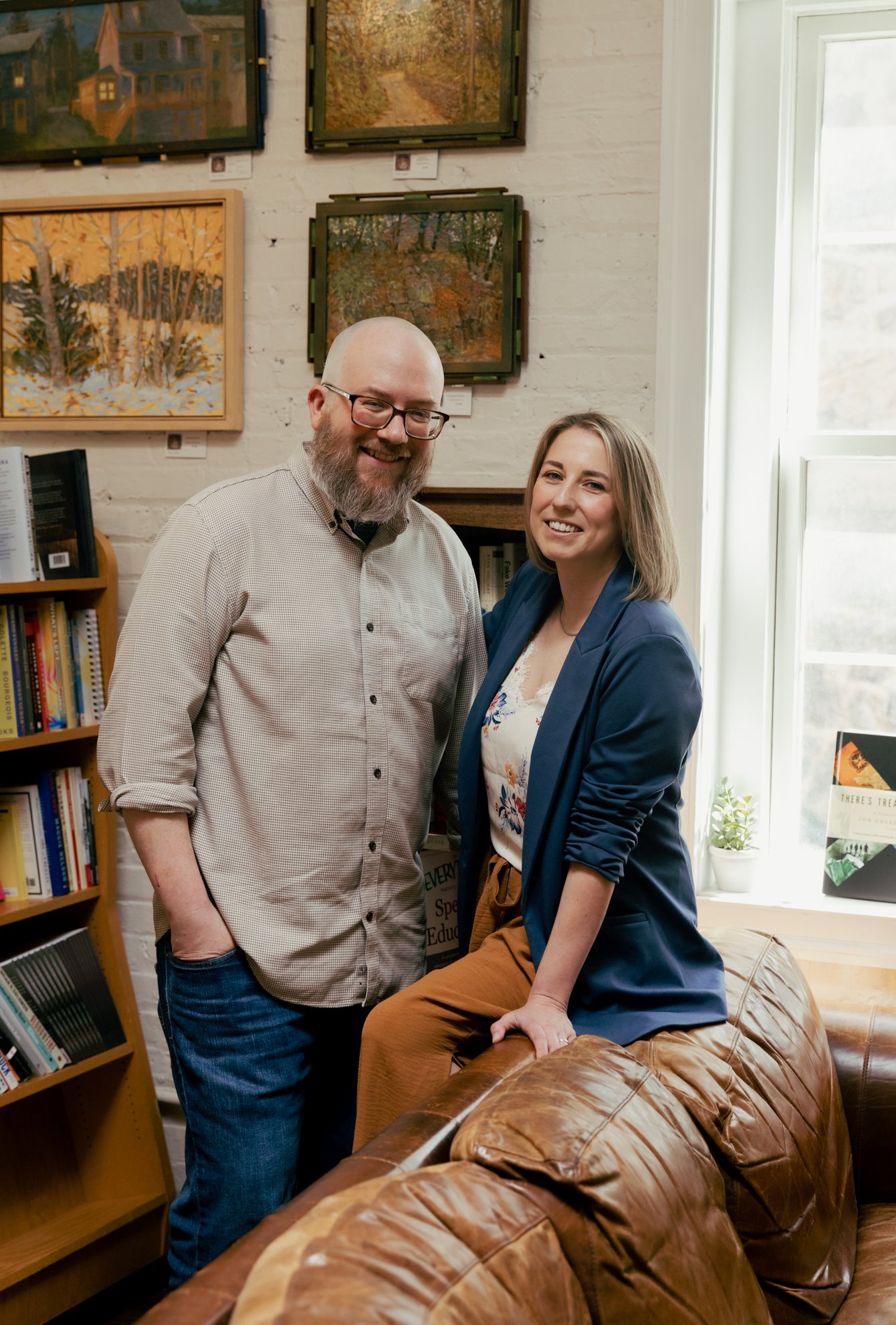 A man with glasses and a beard in a checkered shirt standing next to a woman with shoulder-length blond hair wearing a blue blazer sitting on a leather couch. They are in a room with white brick walls, sunlight coming through a large window, and fram
