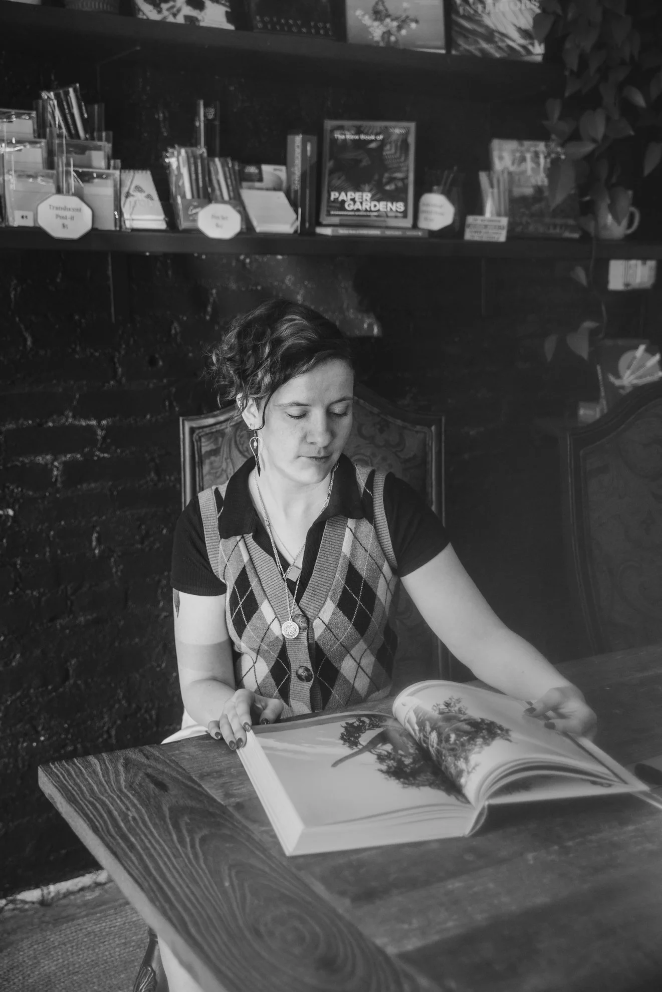 A woman with short curly hair wearing a patterned vest and earrings is sitting at a wooden table, looking at and turning the pages of a large open book or magazine. Behind her, there are bookshelves with books and decorative items in a cozy, possibly