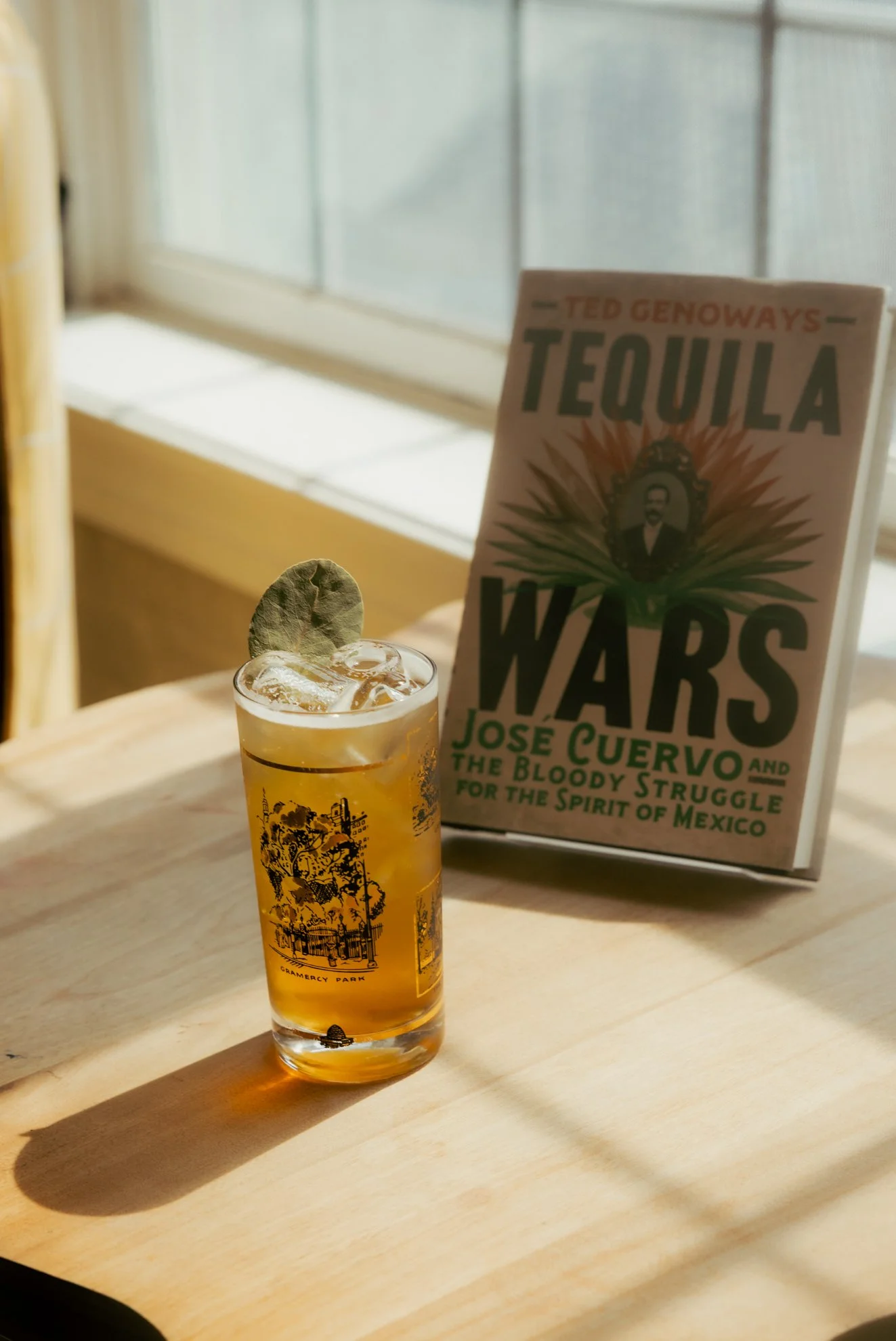 A glass of light amber beer with ice cubes and a dried lime wedge, placed on a wooden table, with a book titled 'Tequila Wars' by Ted Genoways and José Cuervo in the background.