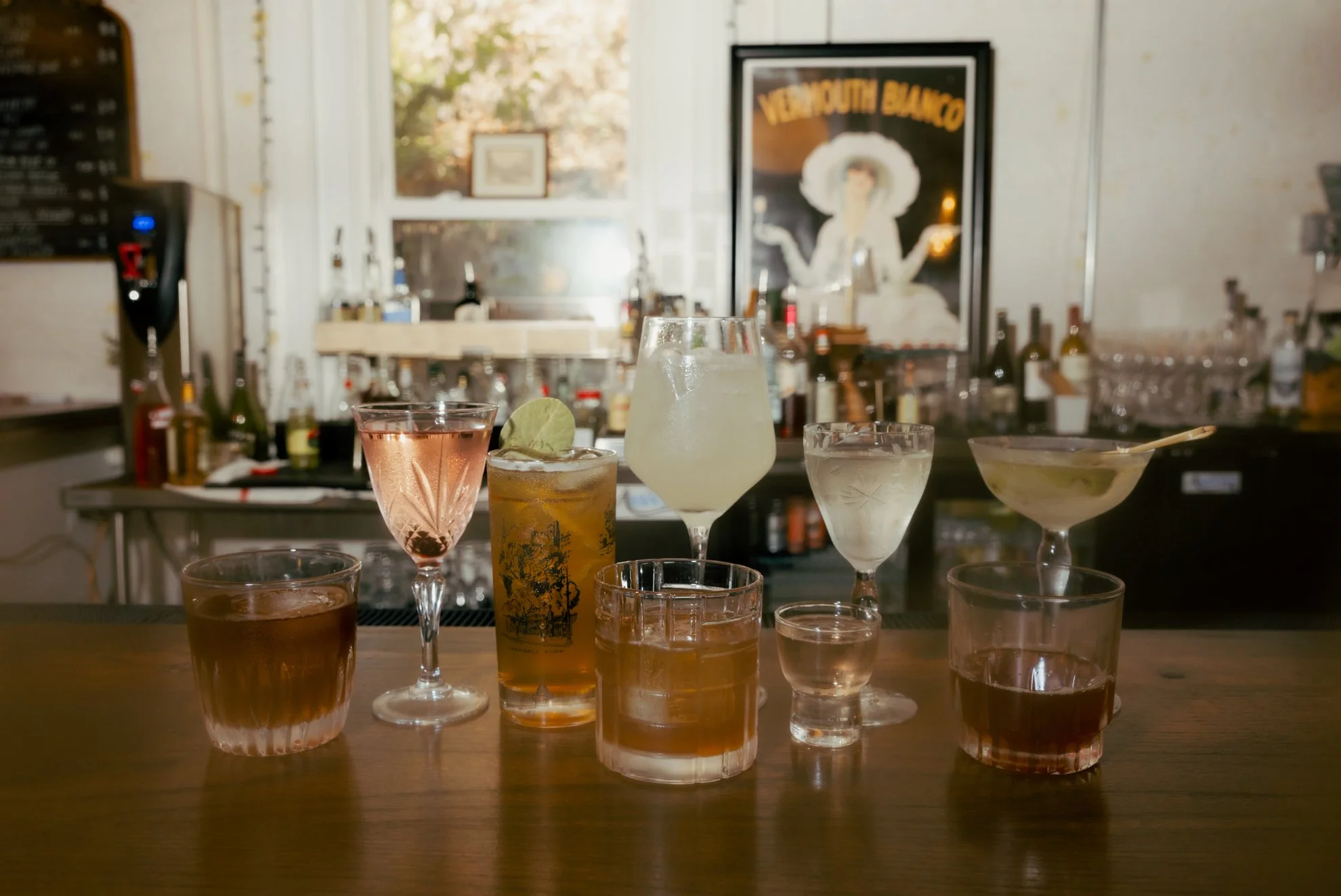 Various cocktails on a bar counter with a blurred bar background, including bottles and a vintage poster.