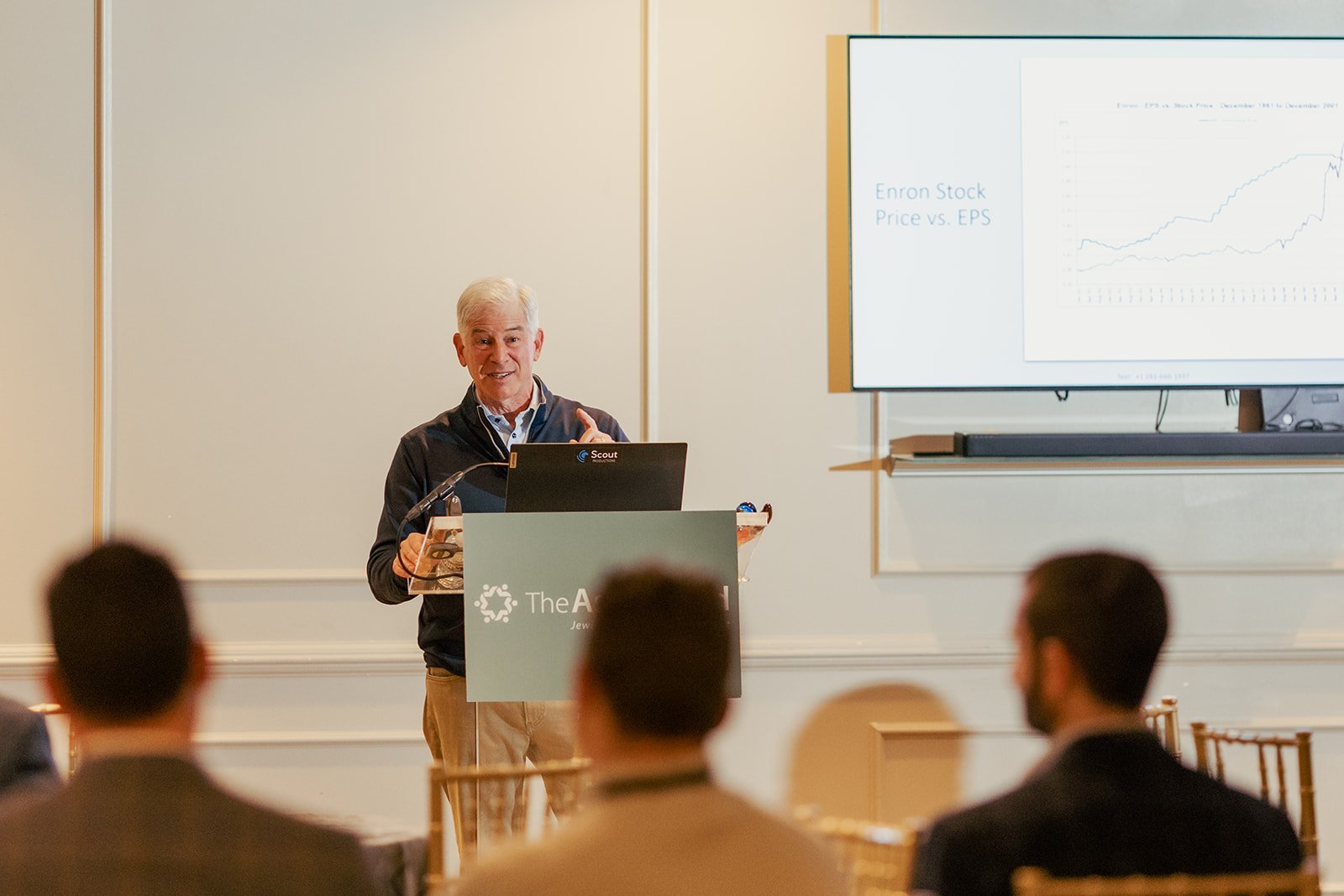 A man giving a presentation to an audience in a conference room, with a slide titled 'Enron Stock Price vs. EPS' displayed on a television screen.
