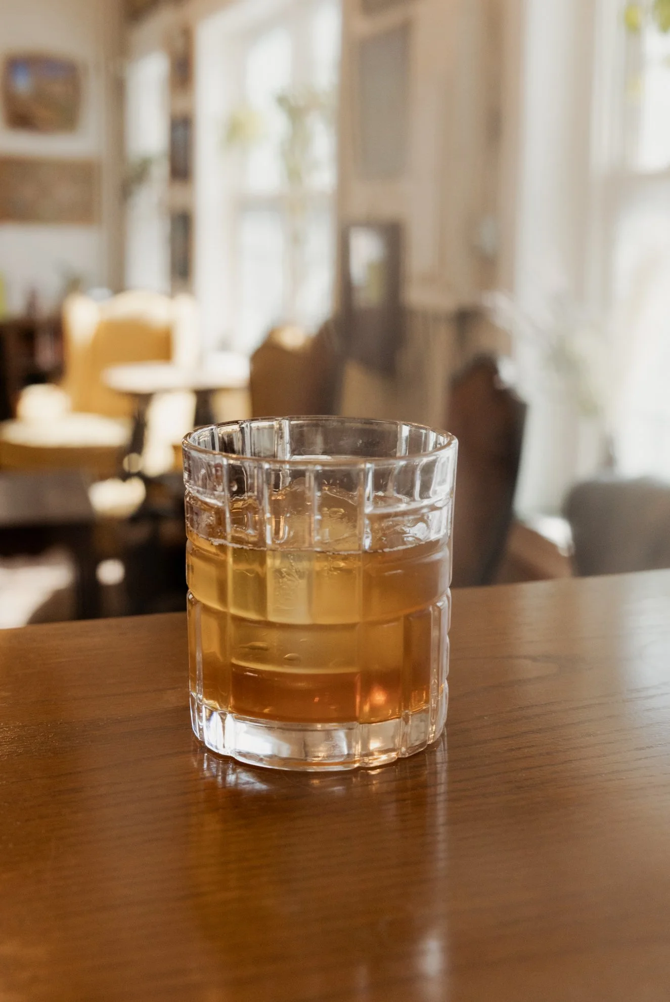 A glass of light-colored beverage on a wooden table in a cozy, well-lit room with blurred furniture and windows in the background.