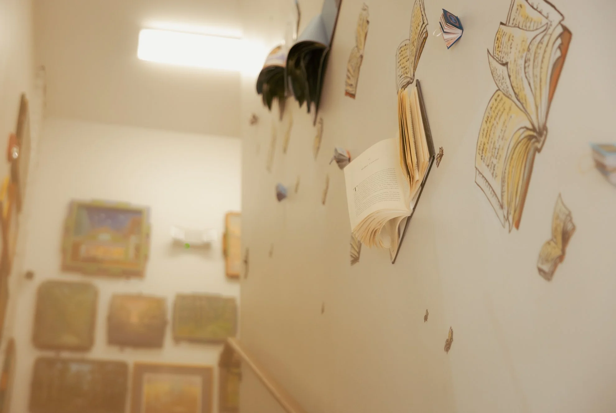 Art display on a wall with open books and folded paper crafts resembling open books, in an art gallery setting with paintings visible in the background.
