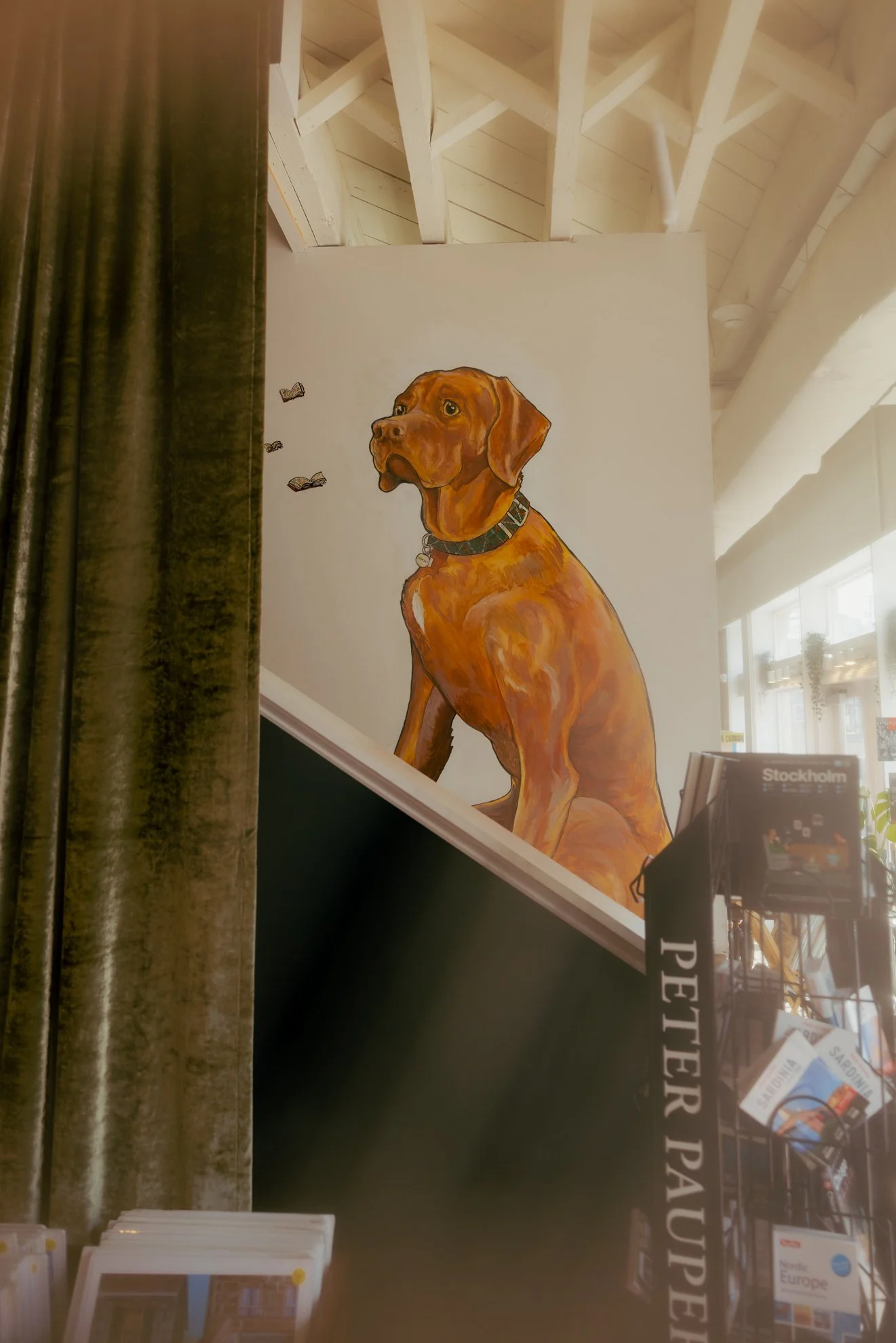 A painted mural of a brown dog sitting, surrounded by flying books on a white wall in an indoor space with a high ceiling and white beams.