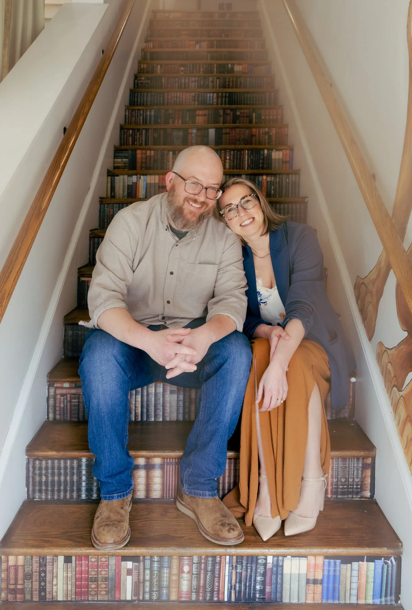 A smiling man and woman sit together on a staircase with bookshelves painted on the risers. They are leaning close, dressed casually and wearing glasses. The staircase has wooden steps with a wood handrail on both sides, and the bookshelves on the ri