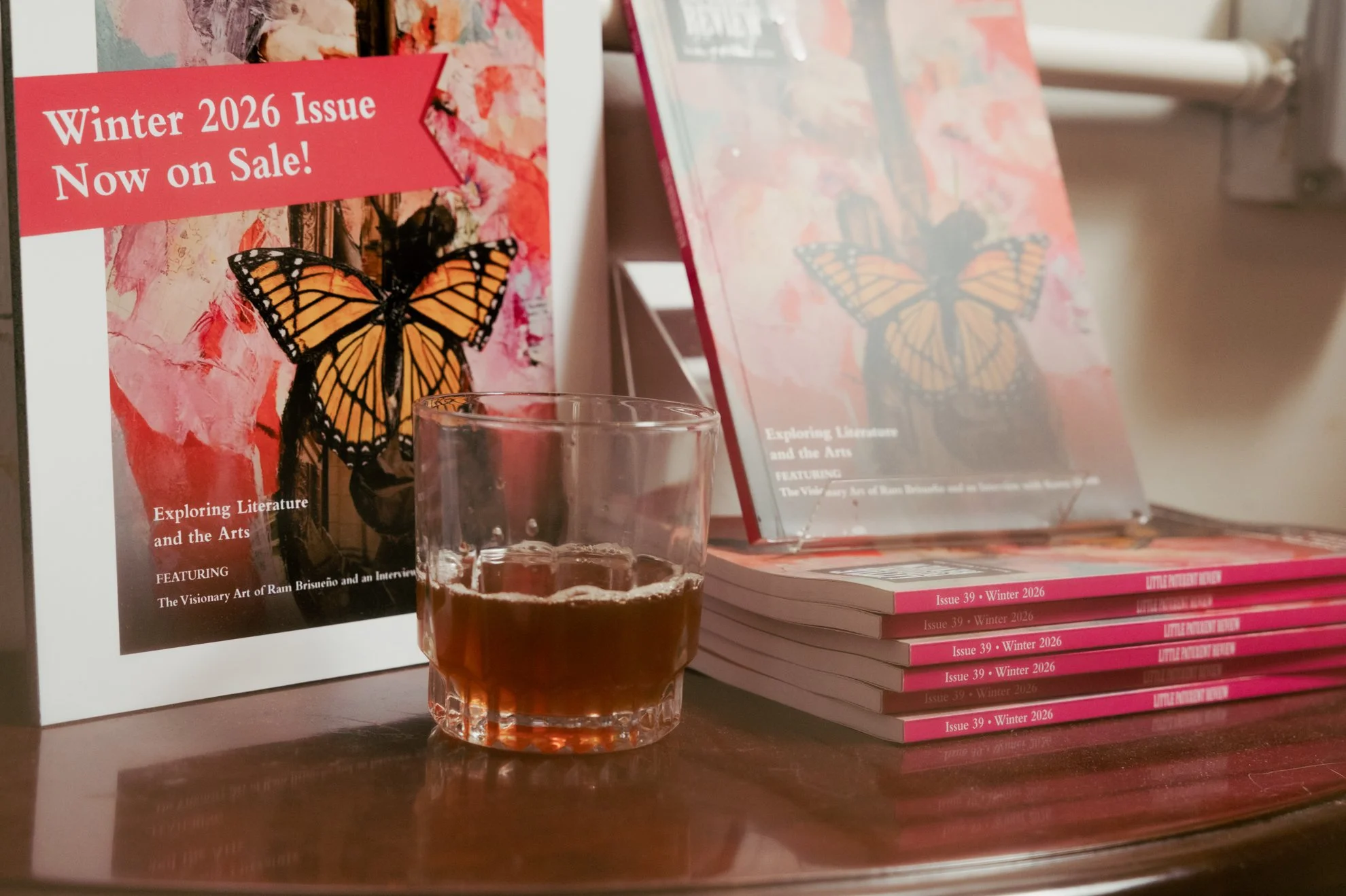 A display of magazines and a glass of iced tea on a wooden surface. The magazines highlight the Winter 2026 issue and feature a butterfly on the cover.