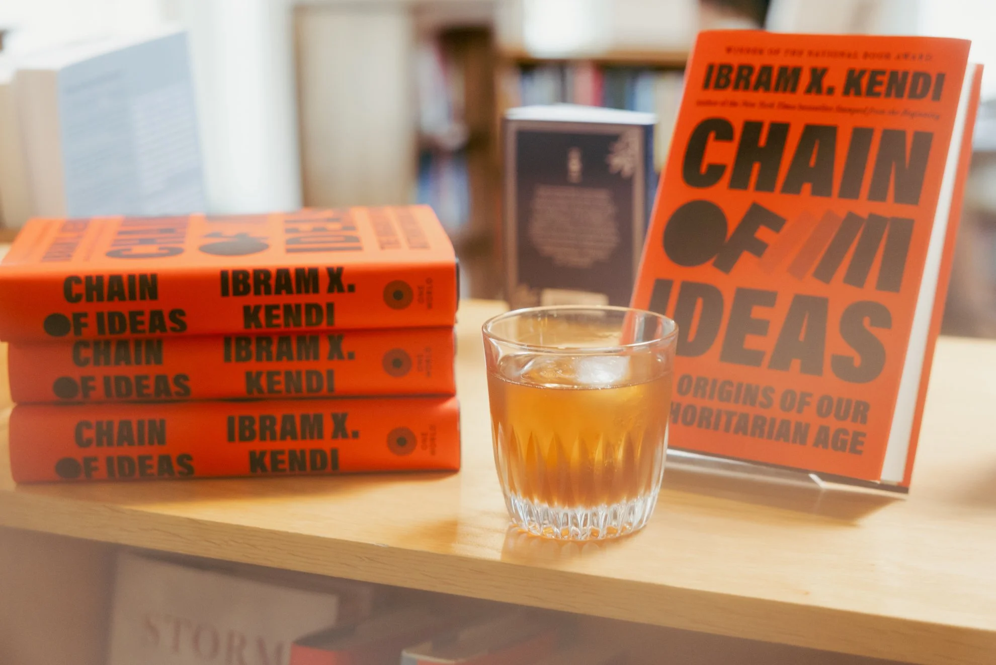 A stack of four orange books titled 'Chain of Ideas' by Ibrahim X. Kendi on a wooden table. Next to the books, there's a clear glass of whiskey with ice and a large orange book titled 'Challenging Ideas' leaning on the second book. In the background,