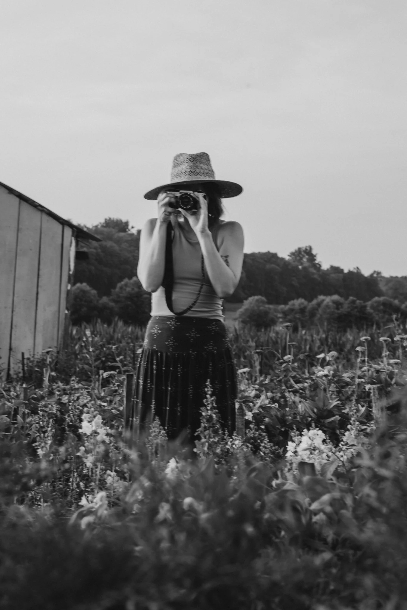 A person wearing a wide-brimmed hat and a sleeveless top holding a camera, standing in a field of flowers with trees and hills in the background, in black and white.