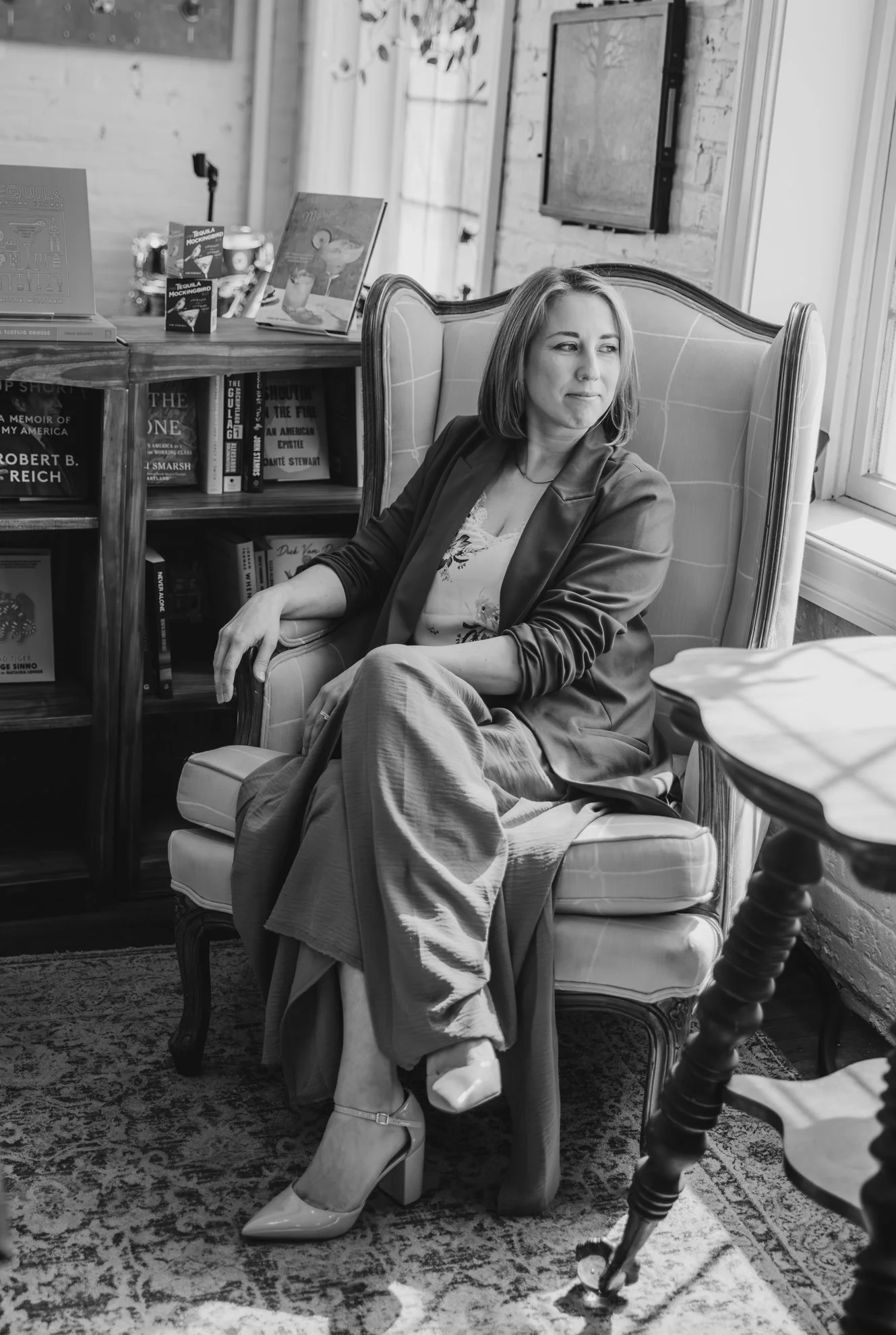 A woman sitting in a high-backed chair in a cozy room with bookshelves and framed pictures, looking to her right.