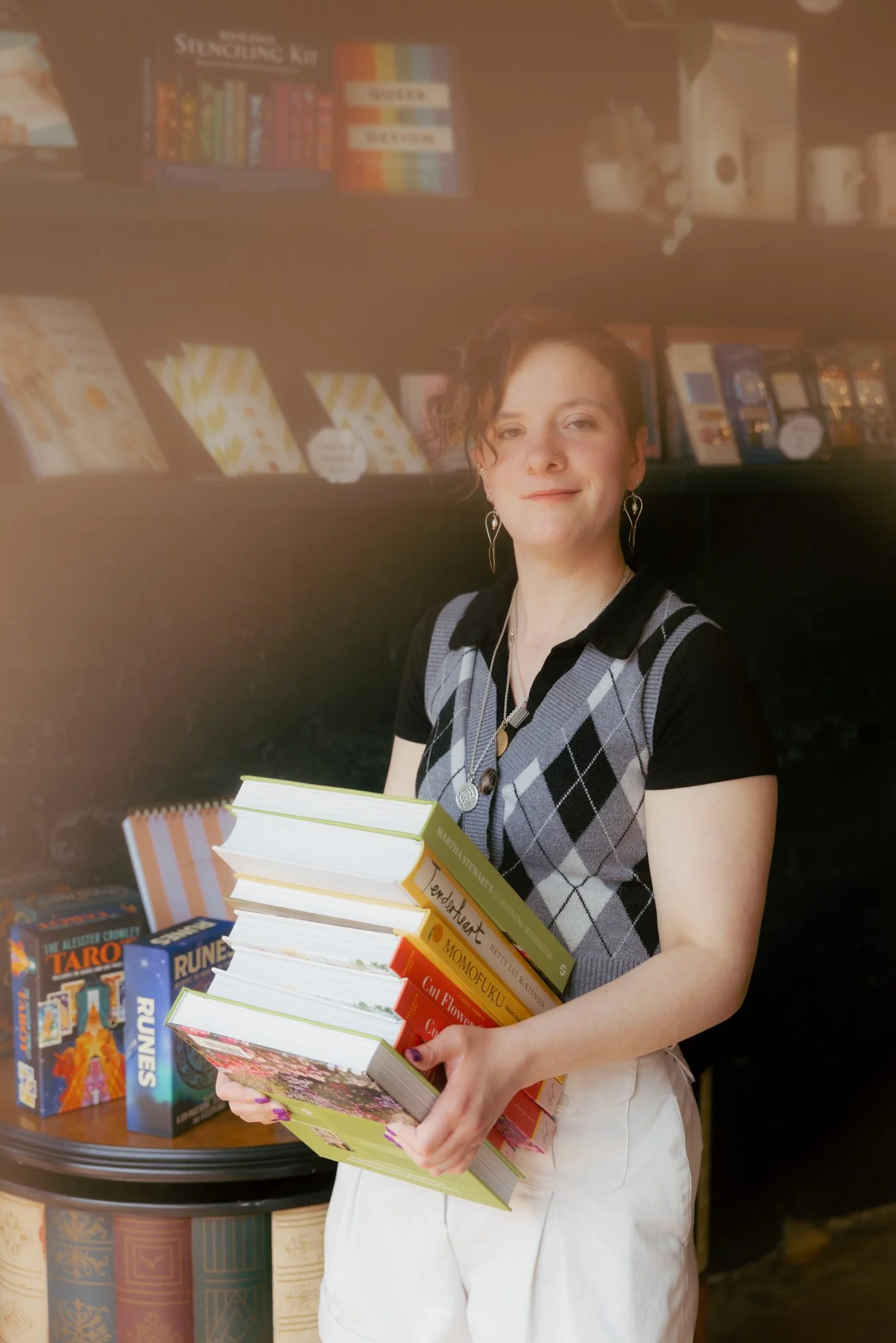 A woman with short wavy hair holding a stack of books in a bookstore. She is wearing earrings and a sleeveless argyle sweater vest over a black shirt. Behind her are bookshelves filled with colorful books and some board games on a table.