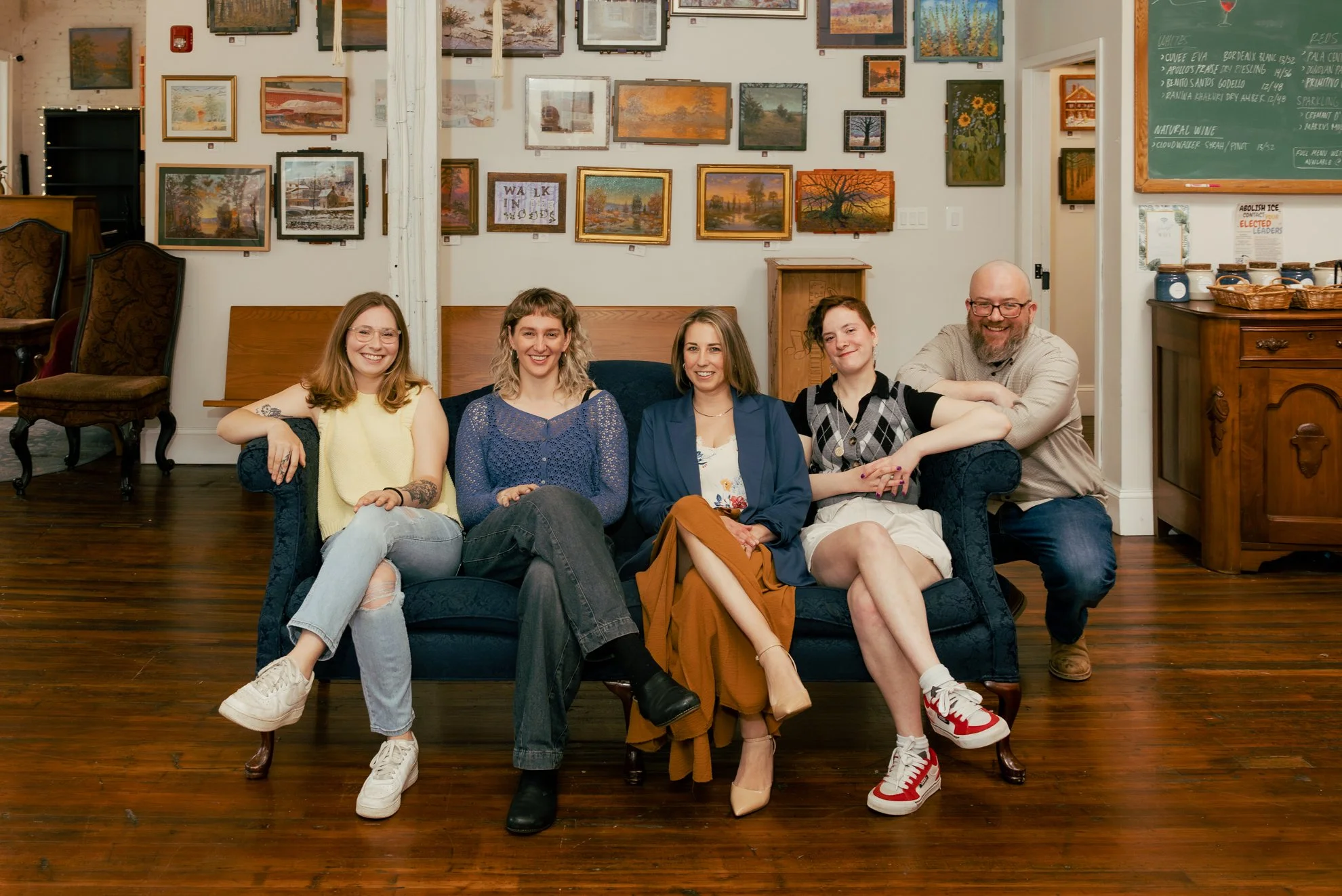 A group of five people sitting on a dark blue sofa in an art gallery or studio, smiling. The background features a wall with numerous framed paintings depicting landscapes and nature scenes. To the side, there is a chalkboard with writing and a woode