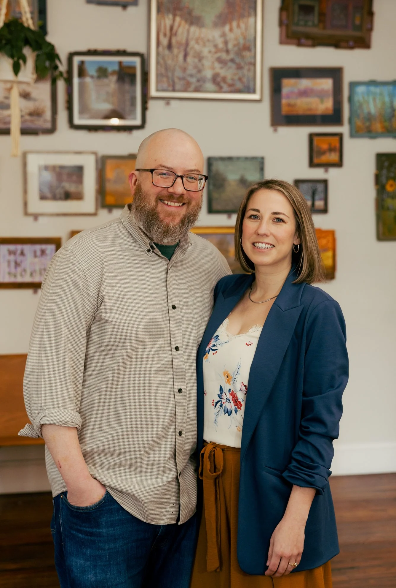 A man and woman standing together, smiling, in an art gallery with framed paintings on the wall behind them.
