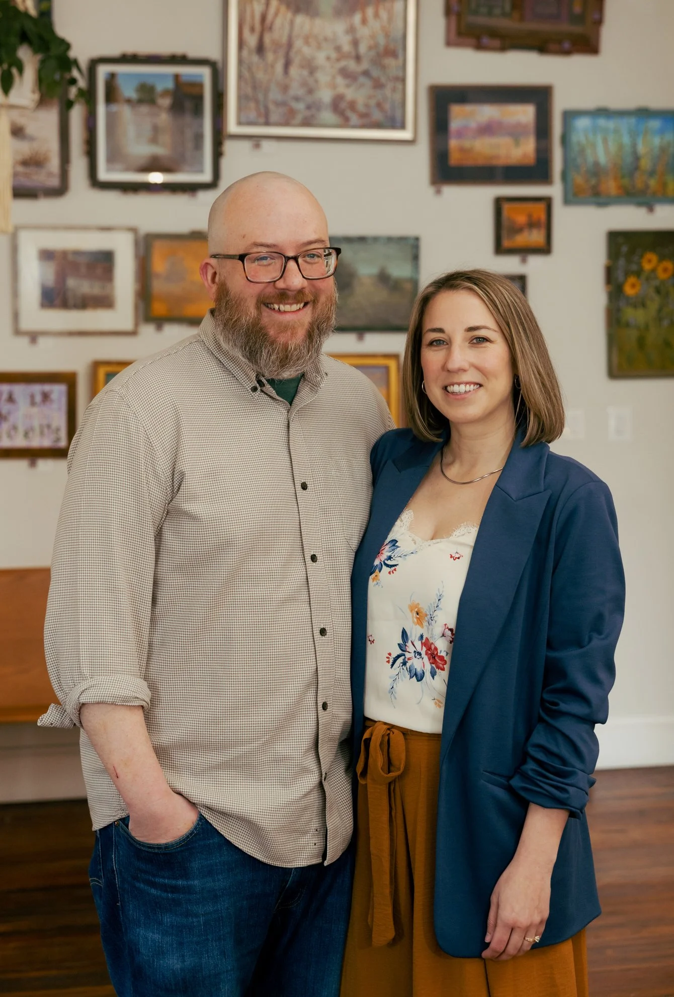 A smiling man with glasses and a beard standing next to a smiling woman with shoulder-length brown hair. They are indoors in an art gallery with framed paintings on the wall behind them.