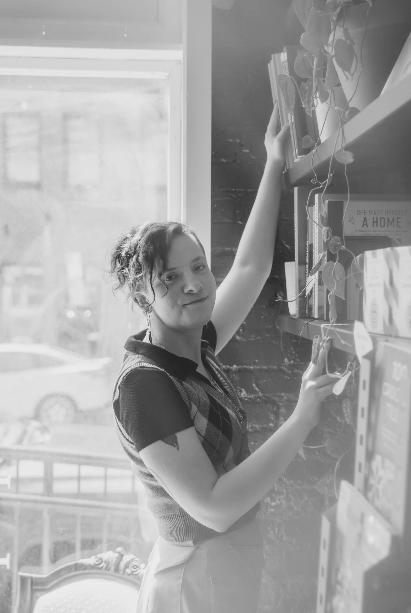 A young woman with curly hair wearing a dark, plaid short-sleeve shirt and light-colored pants, reaching up and holding a book from a shelf with a plant on top, indoors near a window, with a sign that reads 'She made herself a home' in the background