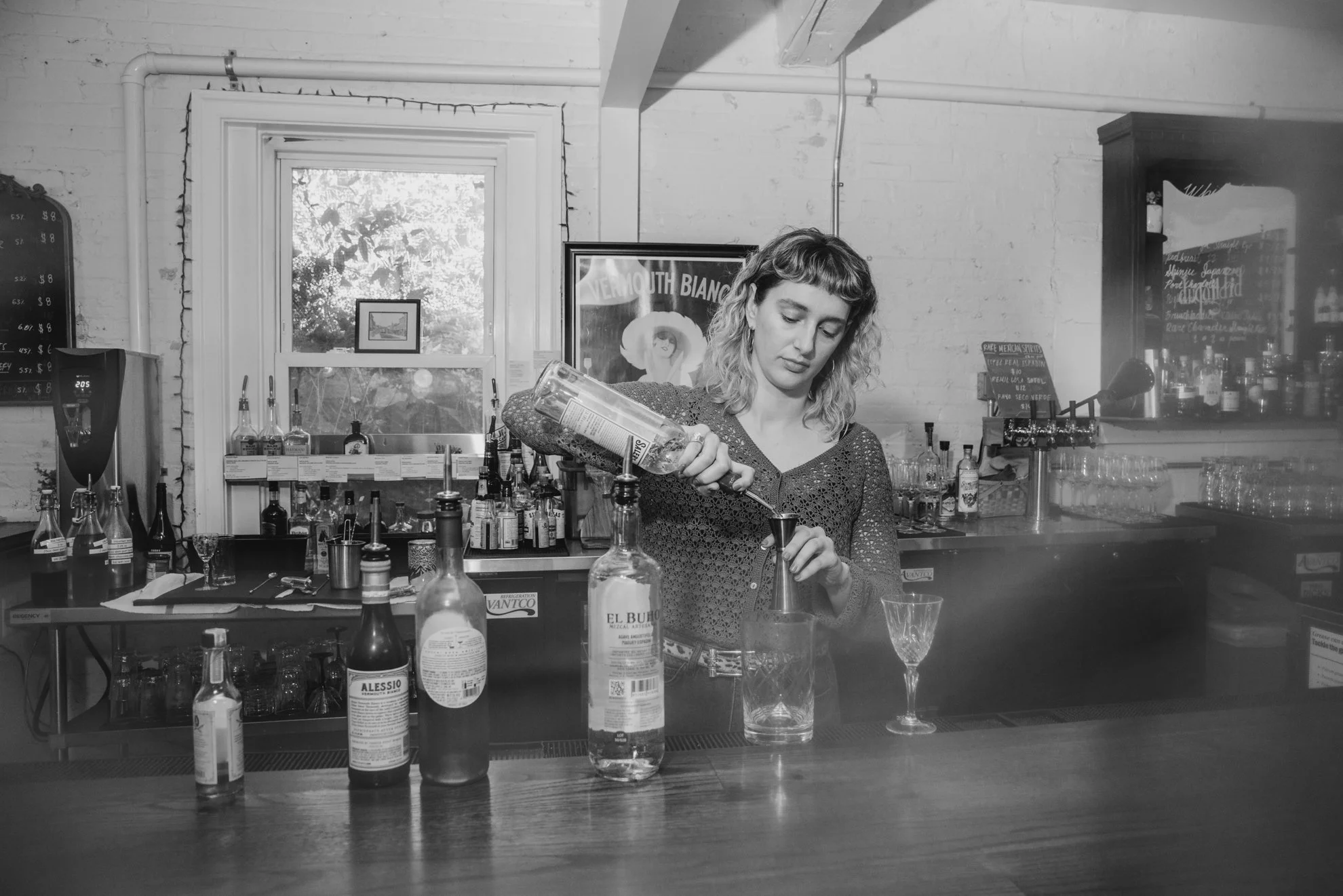A woman working behind a bar, pouring a drink from a bottle into a shaker, with various bottles and bar tools on the counter, in a room with a window and framed posters on the wall.