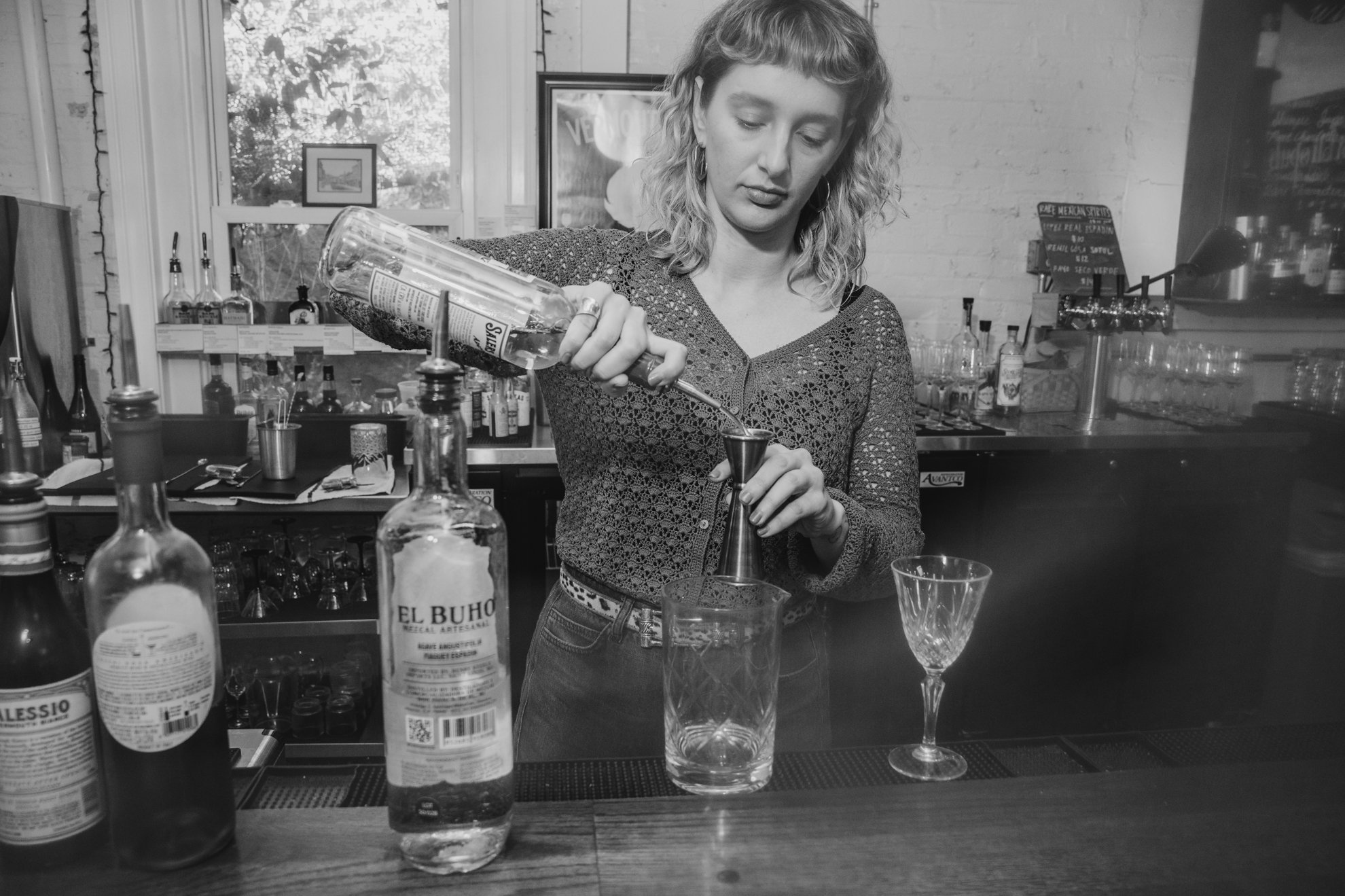 A woman with curly hair and large earrings, wearing a patterned long sleeve shirt, is pouring a clear liquid from a bottle into a metal jigger at a bar. The bar is equipped with various bottles, glasses, and bar tools, with a window in the background