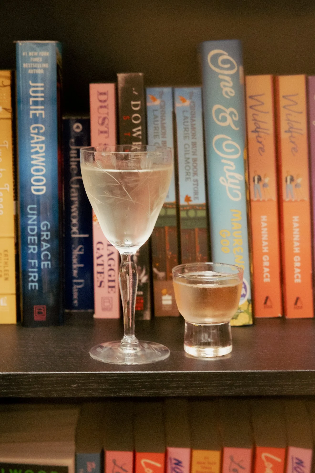A bookshelf with books and two glasses of liquid, one martini glass and one small tumbler, on a dark wooden surface.