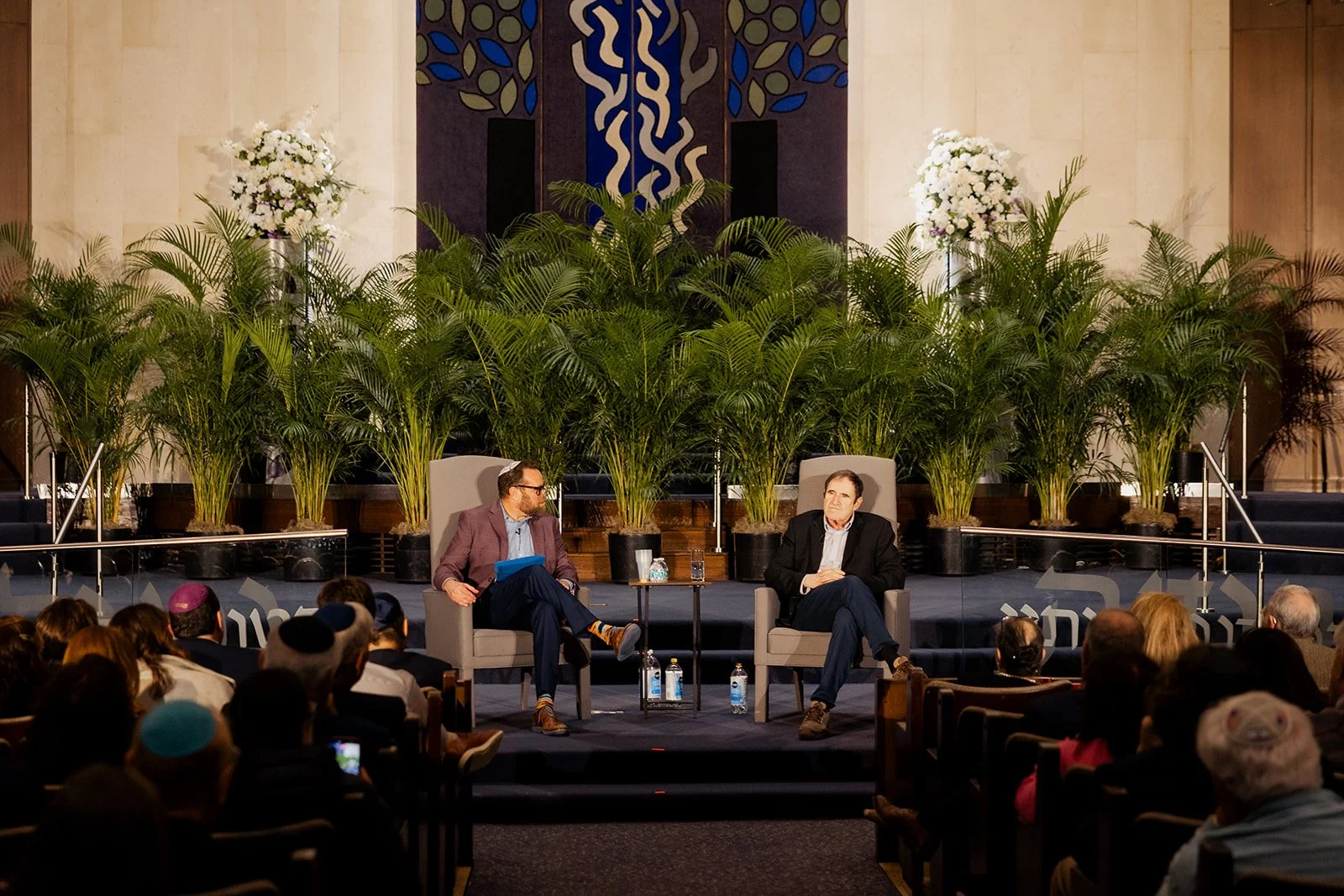 Two men seated in chairs on stage, engaging in a discussion during a conference or event. Behind them is a backdrop with tall green plants and a stained glass wall. Audience members are visible in the foreground.