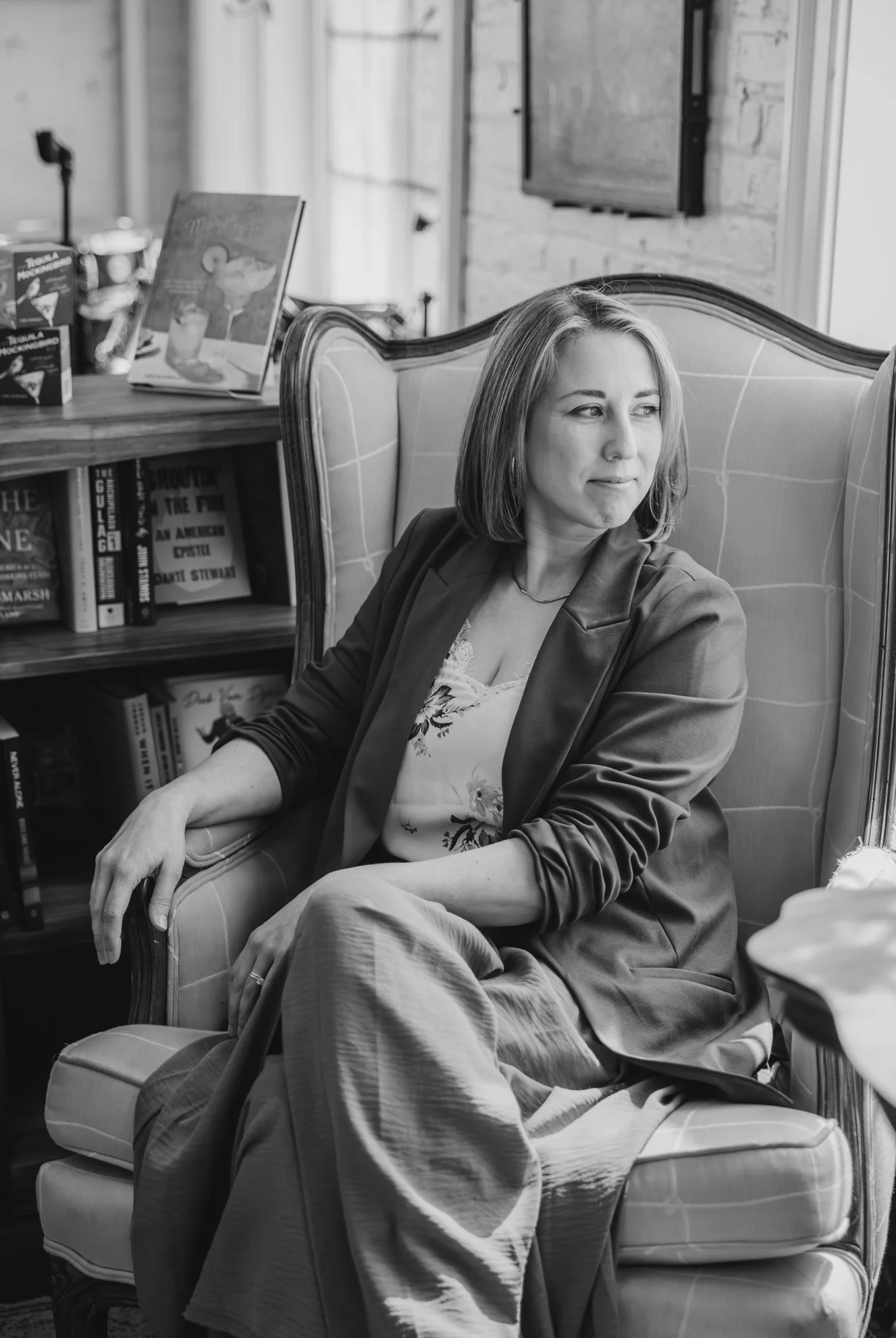 A woman with shoulder-length hair sitting on a patterned armchair in a cozy room, looking to her left. The room has a brick wall, a bookshelf filled with books, and a television mounted on the wall. There are tables with books and objects on them.