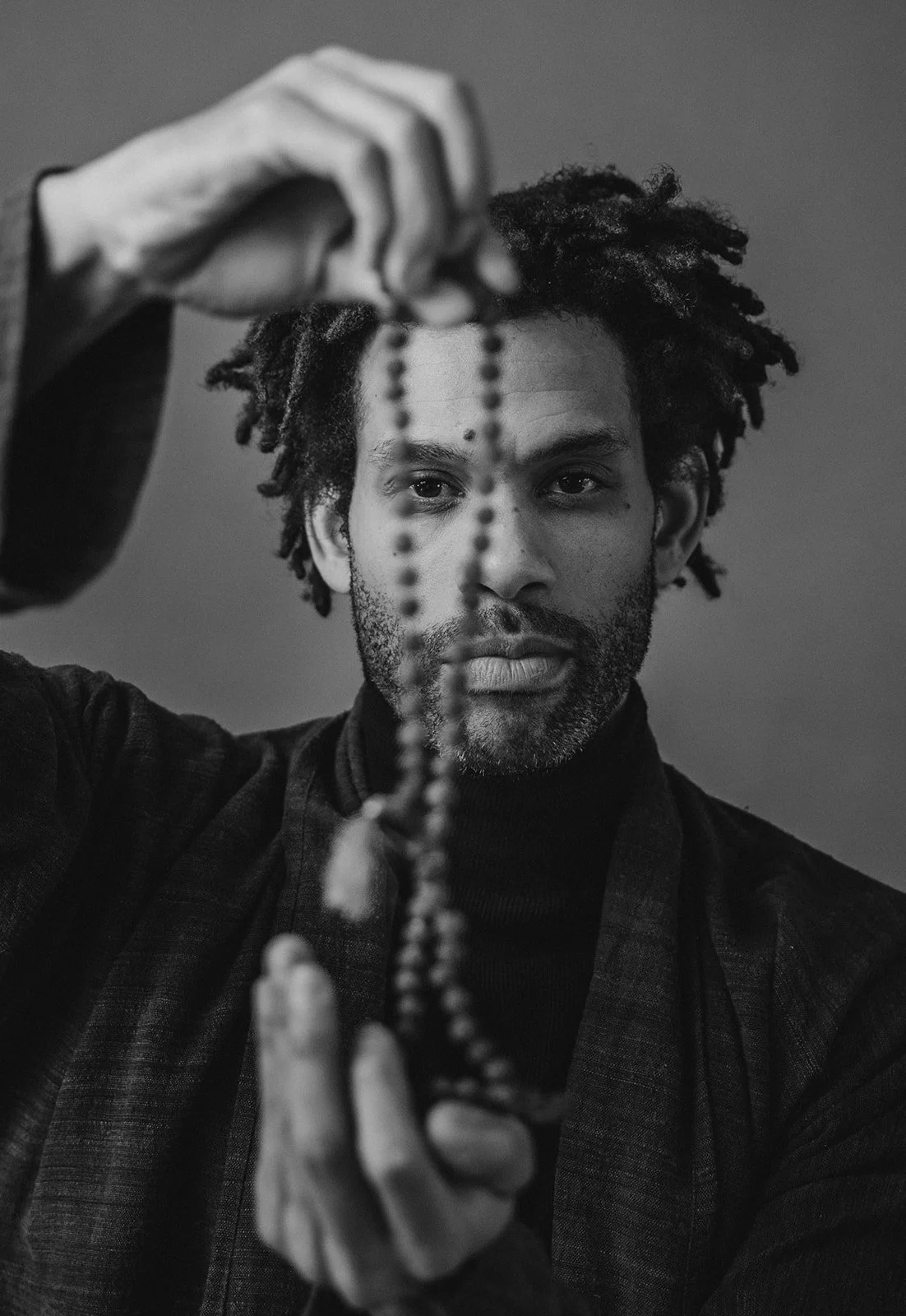 A man with dreadlocks and facial hair holding a beaded necklace in front of his face in black and white.