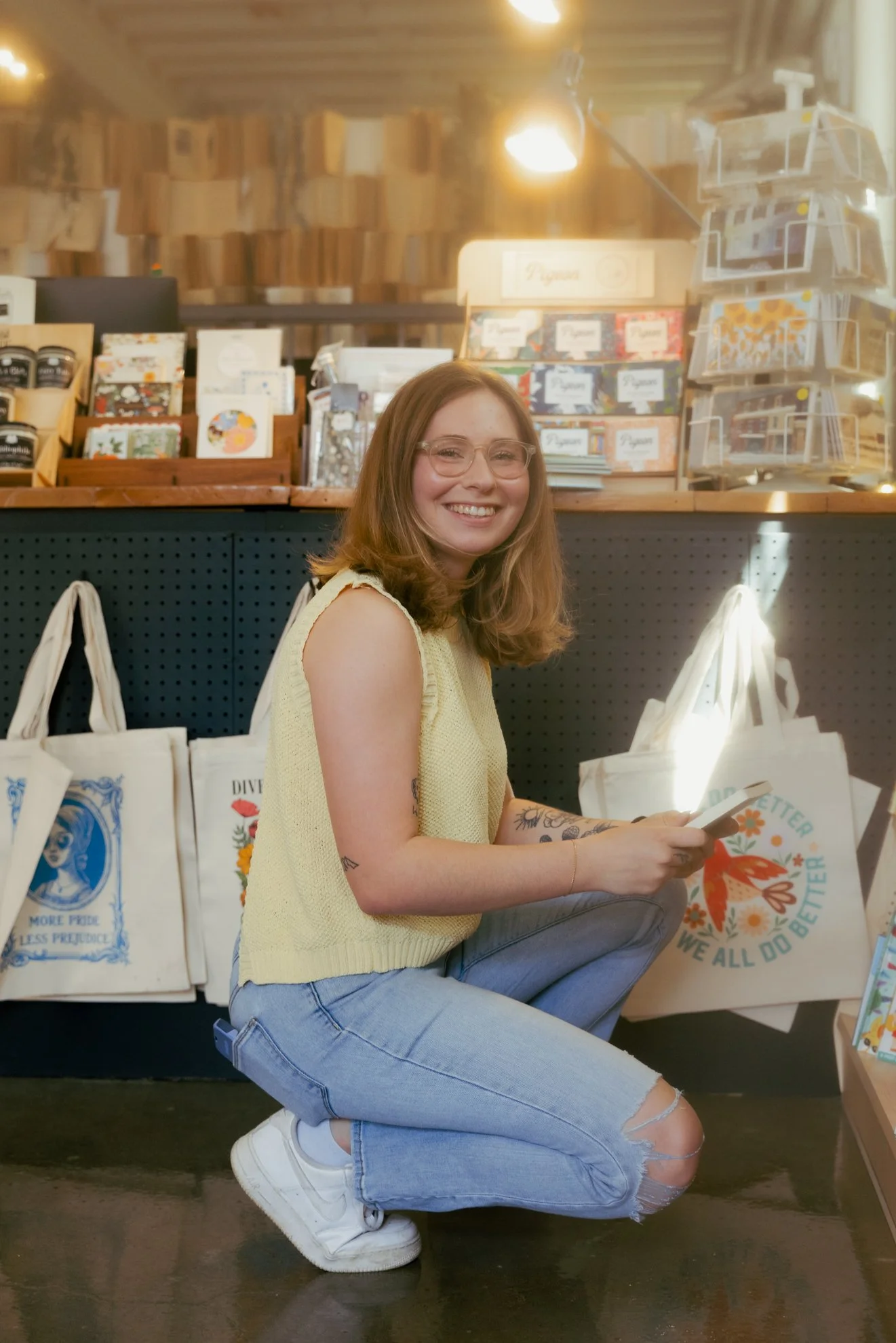 A smiling young woman with glasses and shoulder-length hair kneeling on the floor in front of a display of greeting cards and tote bags at a store. She is wearing a sleeveless yellow top, light blue ripped jeans, and white sneakers. The store has woo