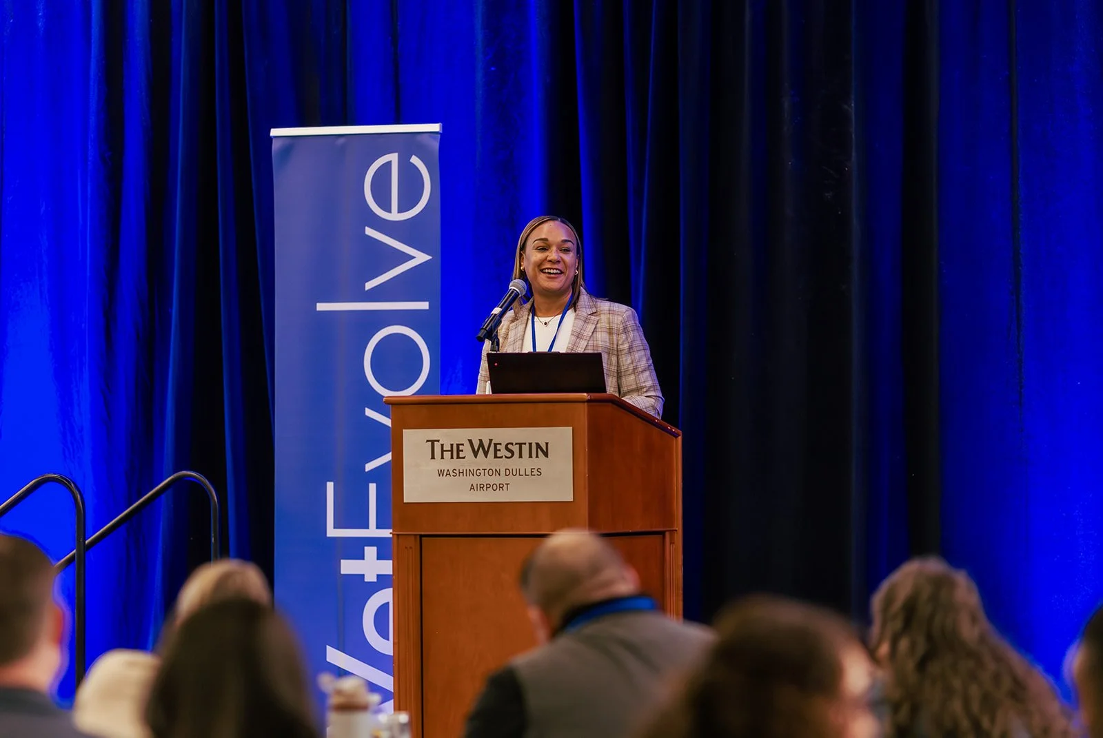 A woman standing at a podium with a microphone, smiling during a conference or presentation at The Westin Washington Dulles Airport, with a blue banner that reads 'Elevate'.