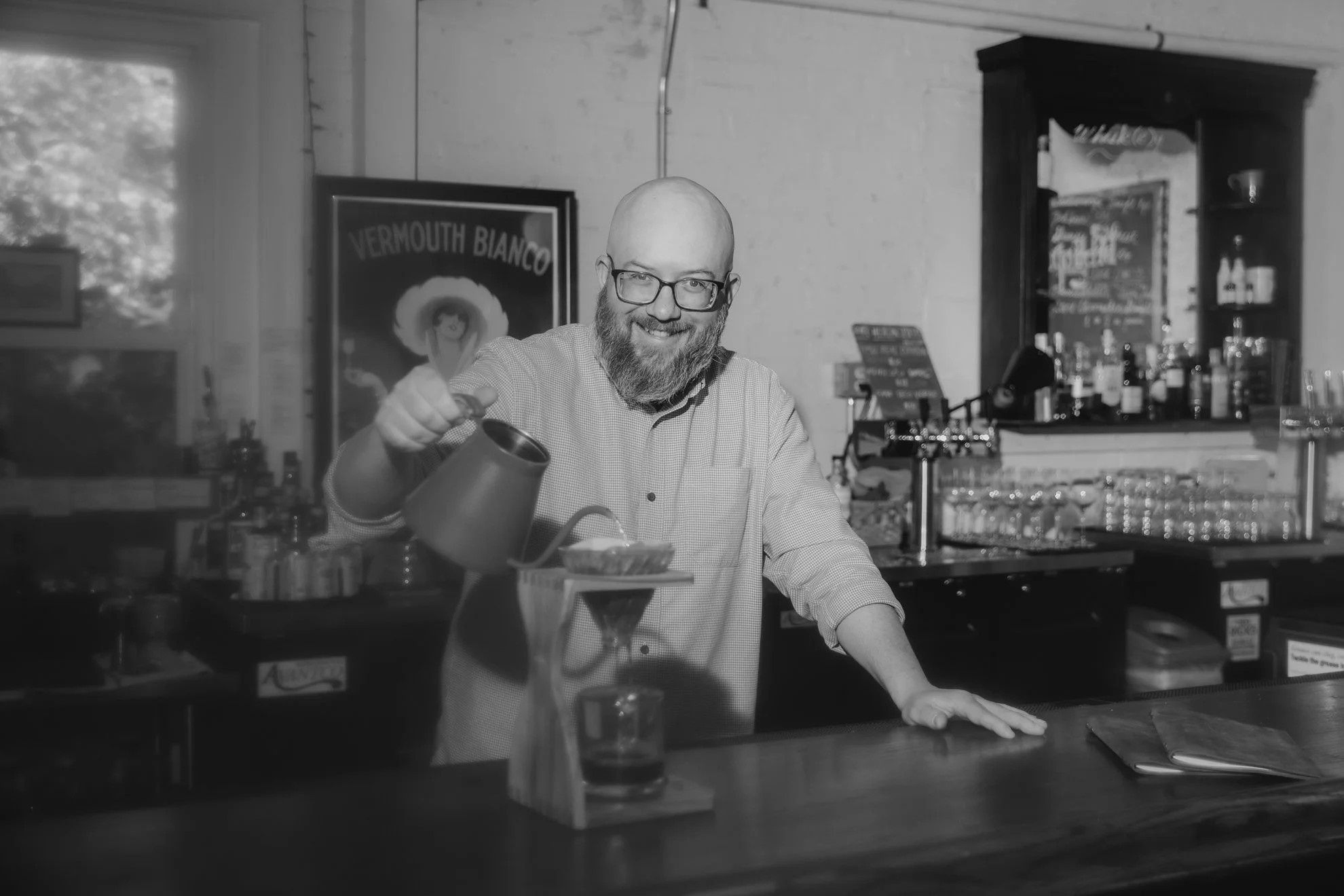 A man with glasses and a beard, smiling, pouring a drink at a bar with bottles and glasses behind him.
