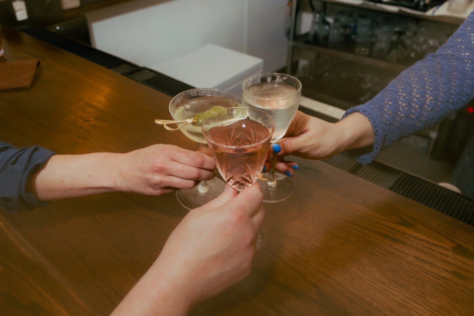 Multiple people clinking glasses with clear and pink-colored beverages in a bar or restaurant setting.