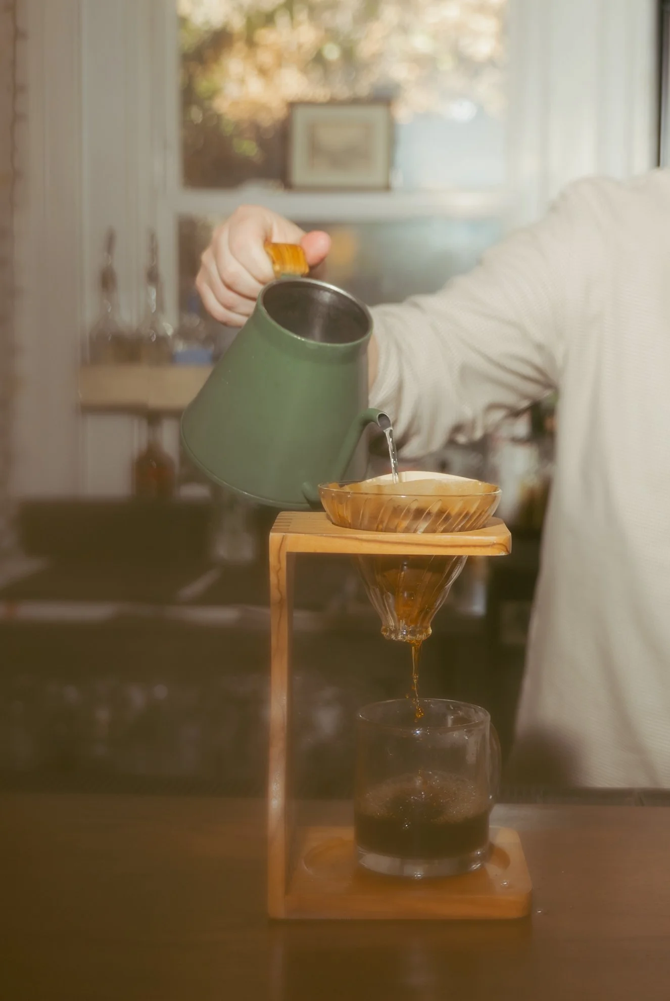 A person pouring coffee through a pour-over coffee maker into a glass on a wooden stand, with a window and framed artwork in the background.