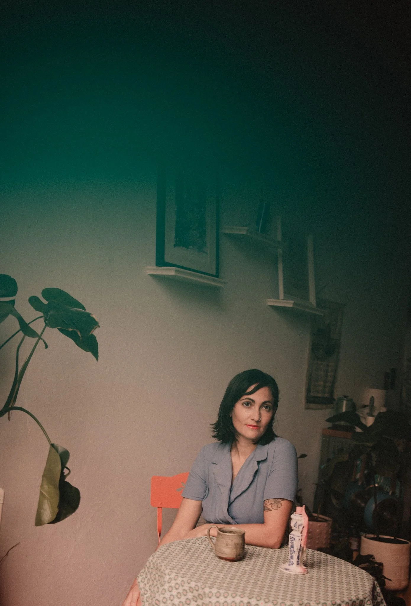A woman with short dark hair and tattoos on her arm sitting at a table with a patterned tablecloth, a mug, and a carton of cream. She is wearing a gray shirt and looking directly at the camera. There are houseplants to her left, framed artwork on the wall behind her, and various objects including a lamp and a bookshelf in the background.