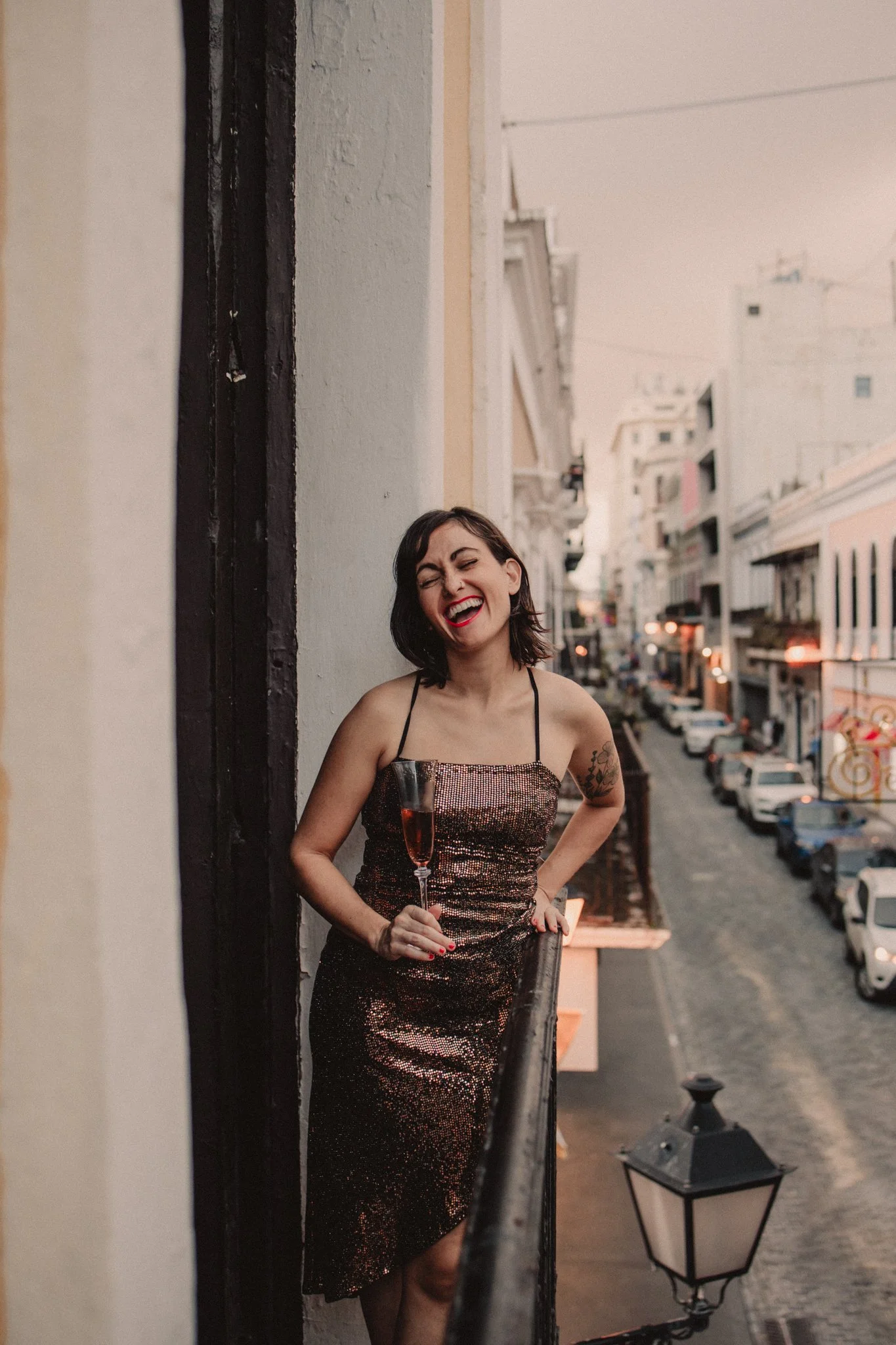 A woman with short dark hair, wearing a sparkly dress, laughing while holding a glass of champagne, standing on a balcony overlooking a city street at dusk