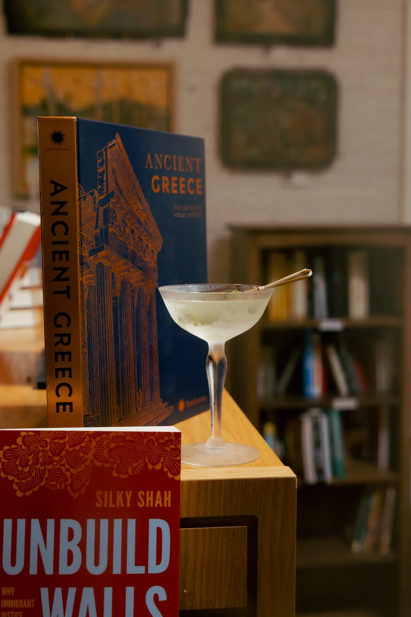 A close-up of a bookshelf with various books and a cocktail glass on top. Prominent books include "Ancient Greece" and "Unbuilding Walls." The background shows framed photos or artwork on a wall.
