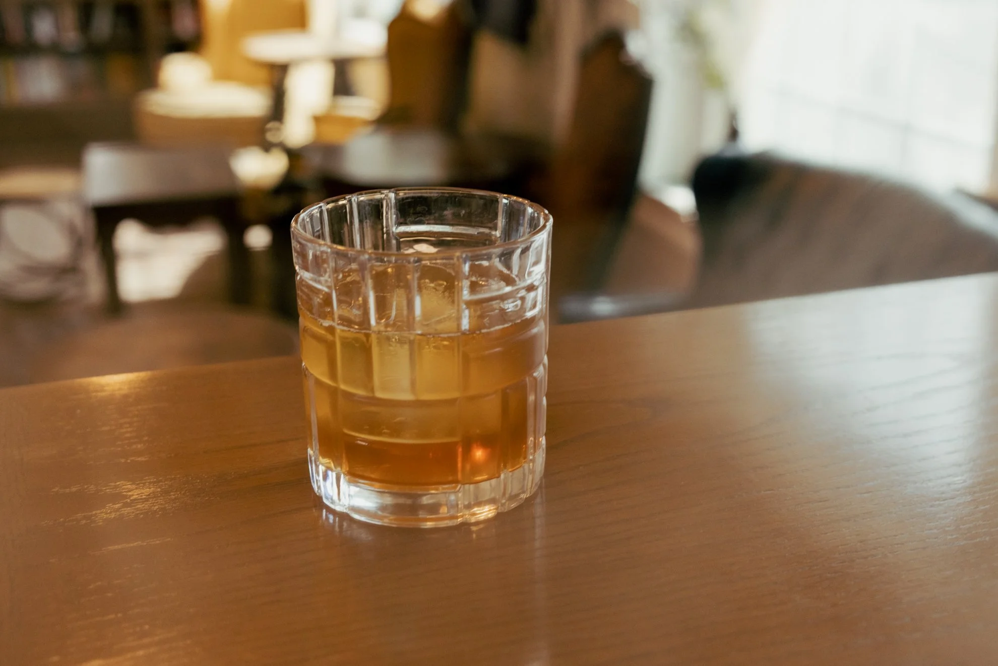 A glass of brownish liquid on a wooden table in a cozy, warmly lit room with blurred background of chairs and a window.