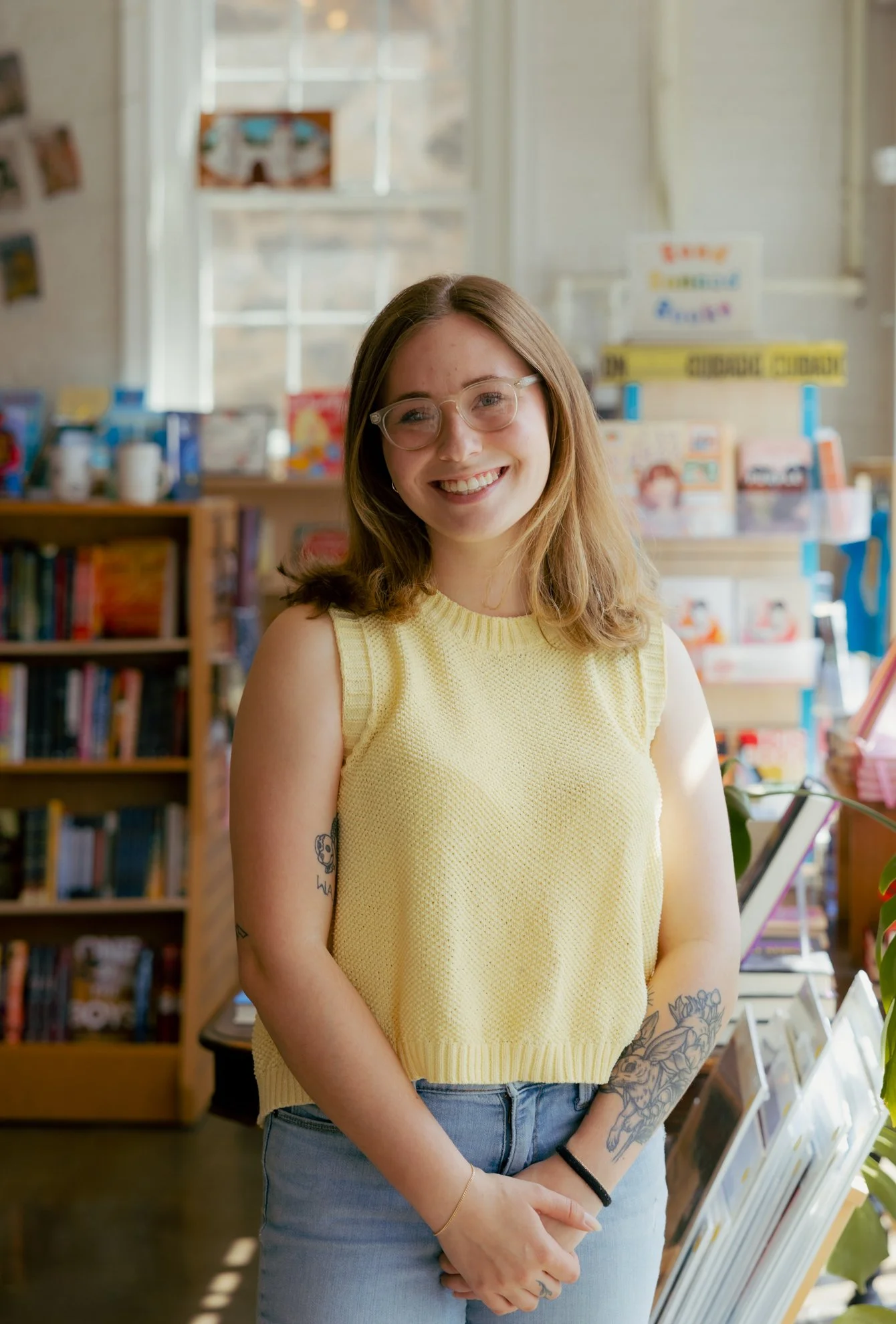 A smiling young woman with glasses and tattoos, wearing a yellow sleeveless top and blue jeans, standing inside a bookstore or library with bookshelves and displays in the background.