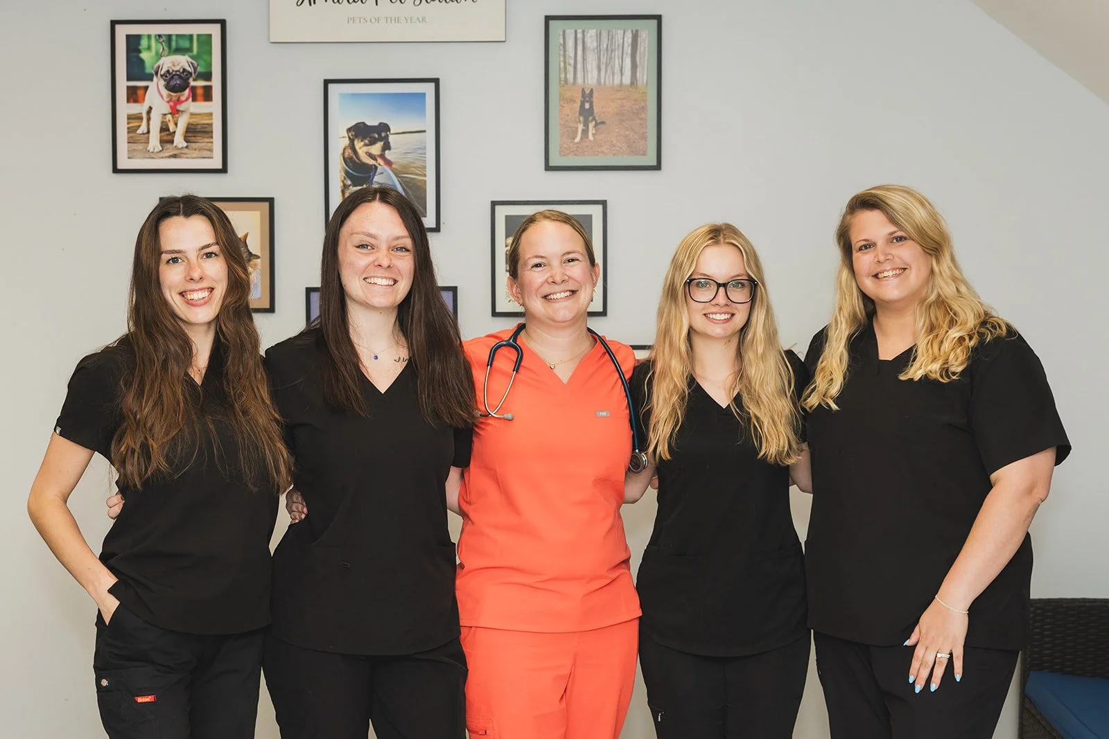 Group of five female veterinary professionals standing together in front of a wall decorated with dog-themed framed photos, all smiling and wearing scrubs.