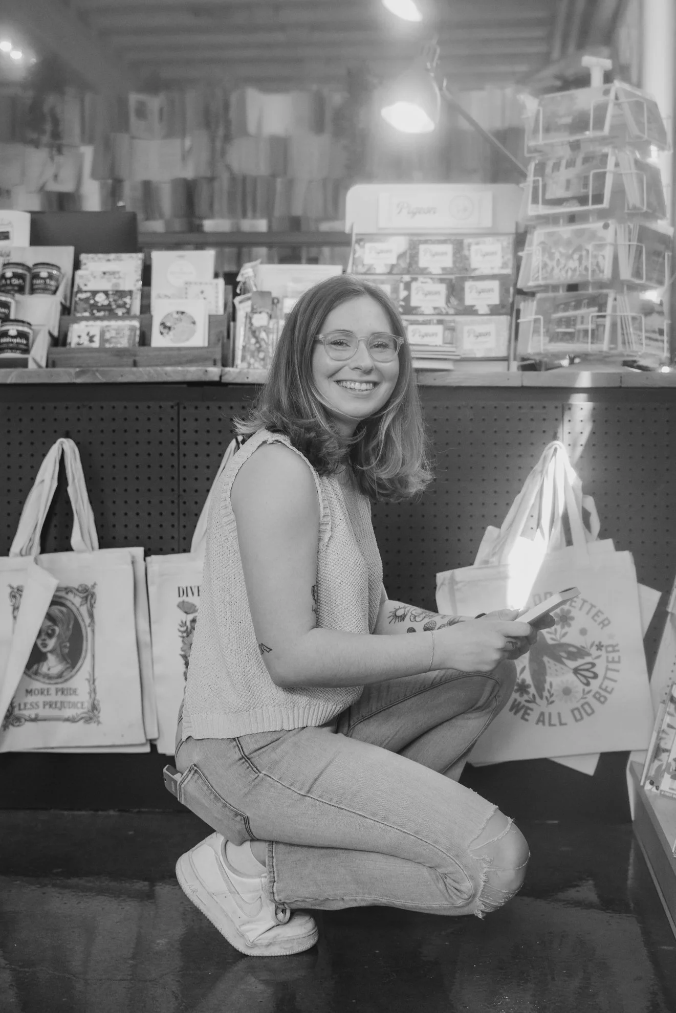 A young woman with glasses, smiling, crouching in front of a display of greeting cards and tote bags at a store. She has shoulder-length hair, is wearing a sleeveless sweater and ripped jeans, holding a smartphone. The background shows shelves with m