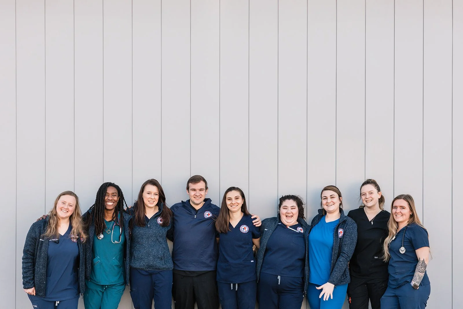 Group of nine healthcare professionals standing together outside, smiling, in front of a plain gray wall. They are wearing scrubs and jackets with medical emblems, some with stethoscopes around their necks.